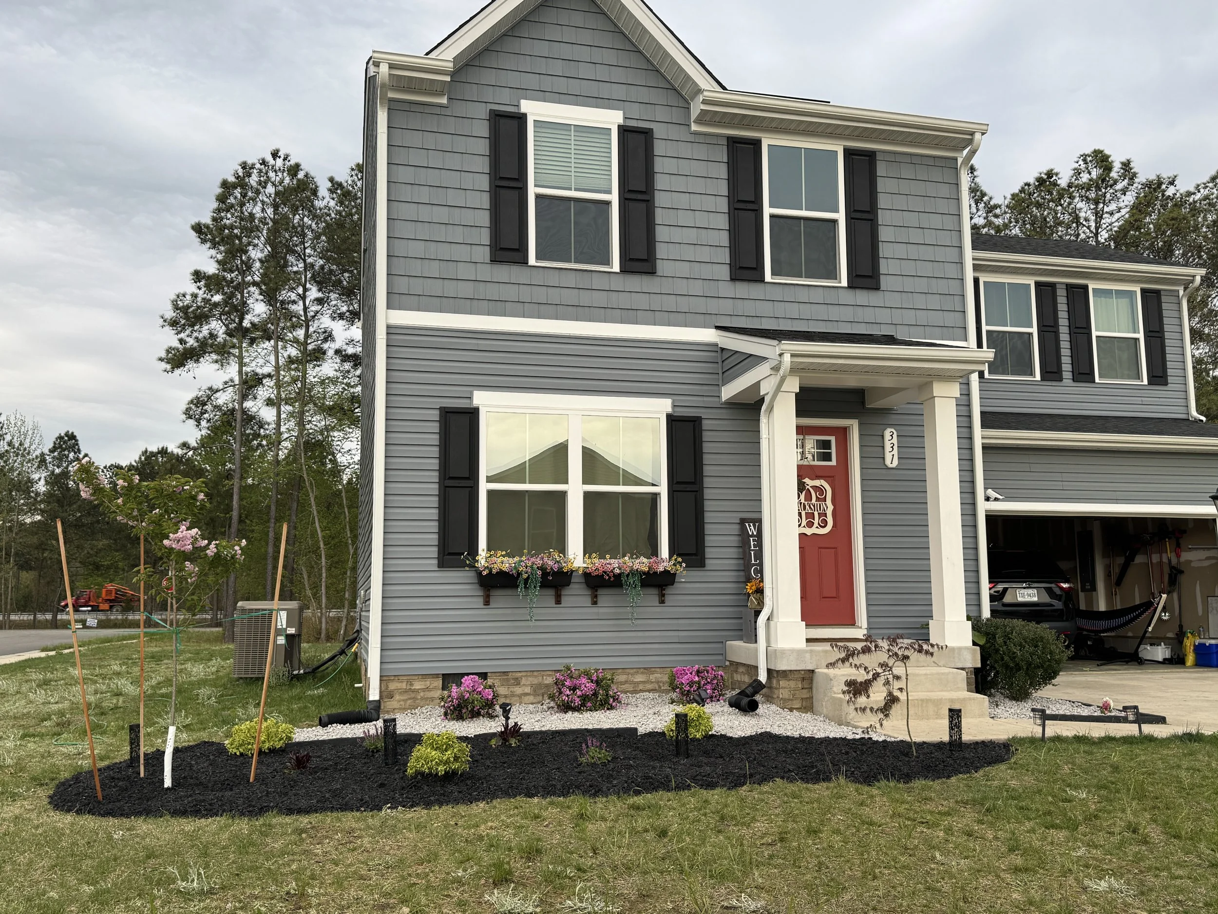 Front view of a two-story gray house with black window shutters, a red front door, and a flower bed with blooming plants, surrounded by a grassy lawn.