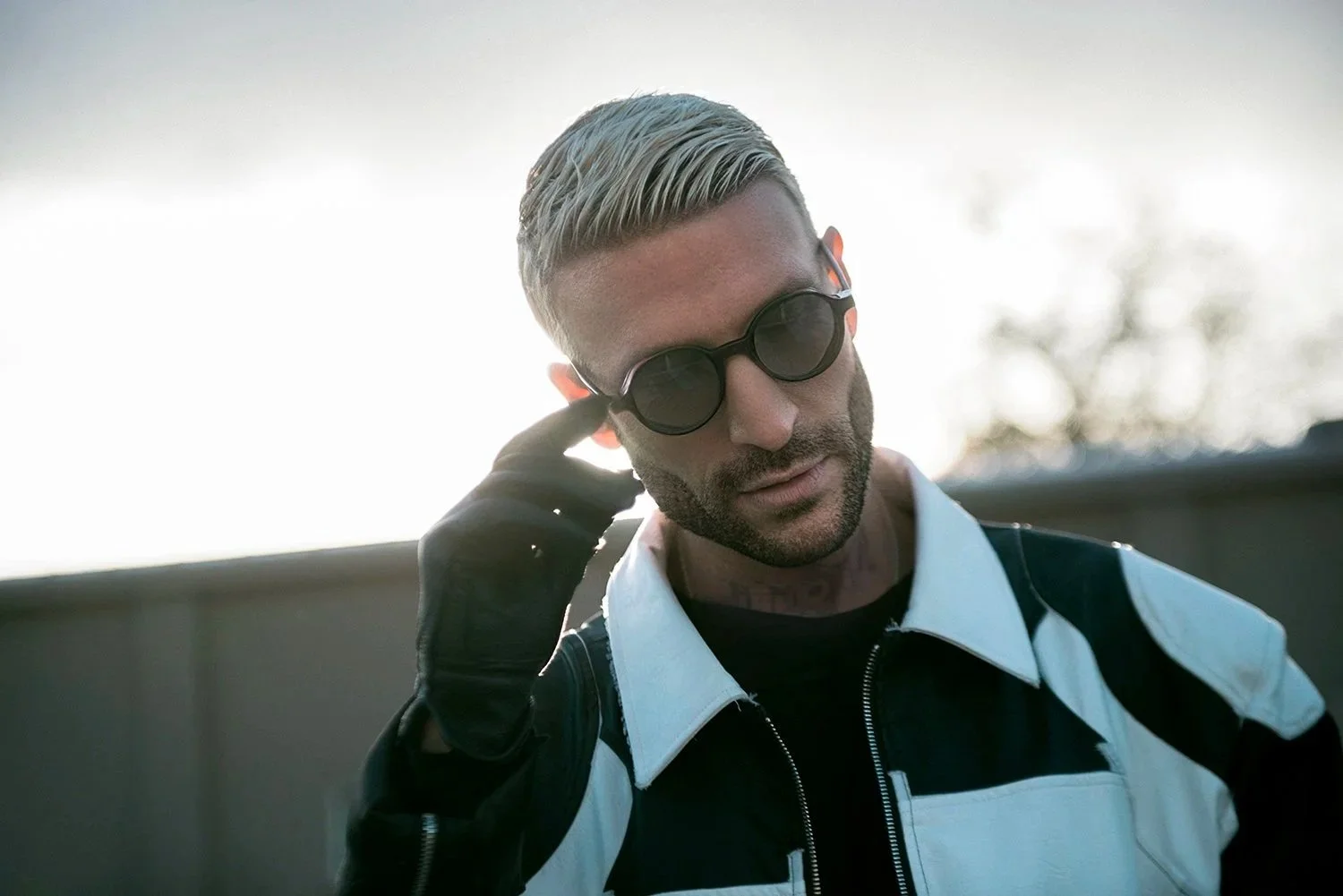 A man with platinum blonde hair, wearing round black sunglasses, black gloves, a black and white motorcycle jacket, and a black shirt, standing outdoors with a bright sky and trees in the background.