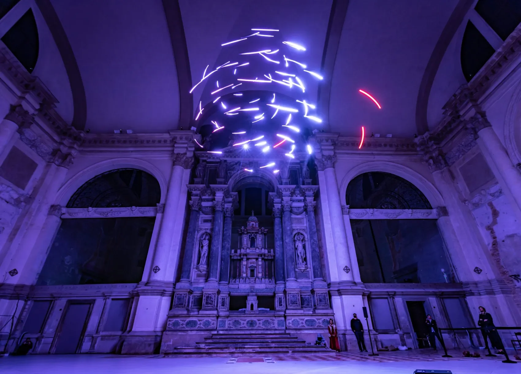 An interior with classical architecture featuring ornate columns and arches, illuminated by purple and blue lighting, with abstract neon light art in the ceiling and people standing near the walls. Don Diablo, Drift.