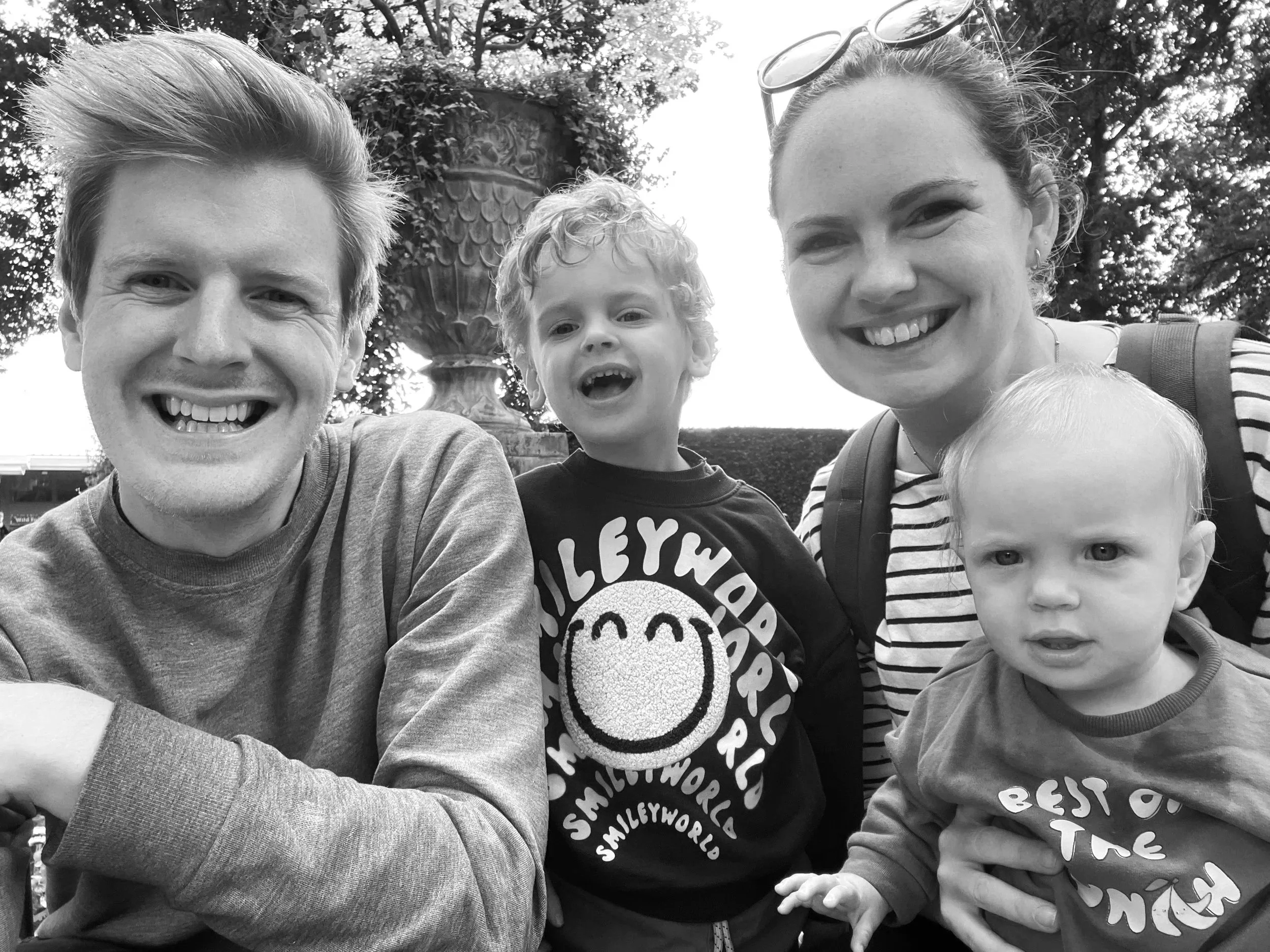 A smiling family of four taking a selfie outdoors. The father and mother are smiling with two young children, one boy and one girl, in front of a large decorative fountain with trees in the background.