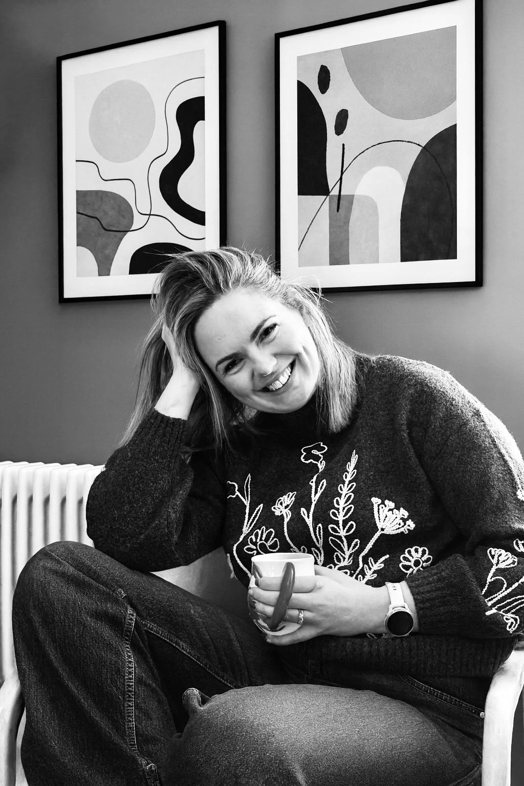 A woman with shoulder-length hair, smiling and holding a mug, sitting on a chair in front of two abstract art pieces on the wall.