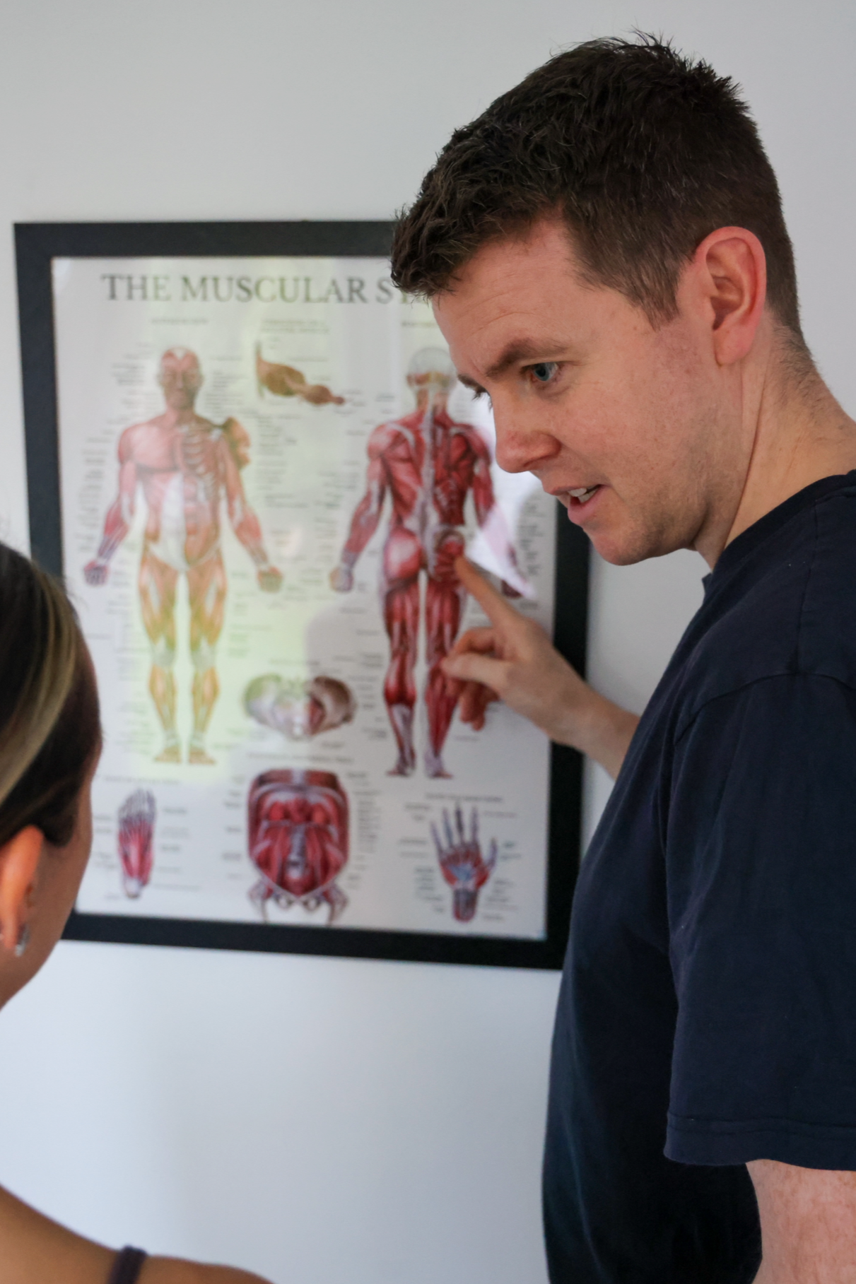 A man in a dark shirt pointing at a muscular human anatomy poster while talking to a woman.