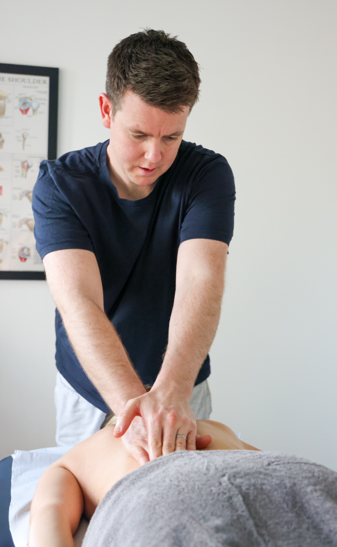 A man giving a back massage to a person lying face down on a massage table in a clinic room with a medical chart on the wall.