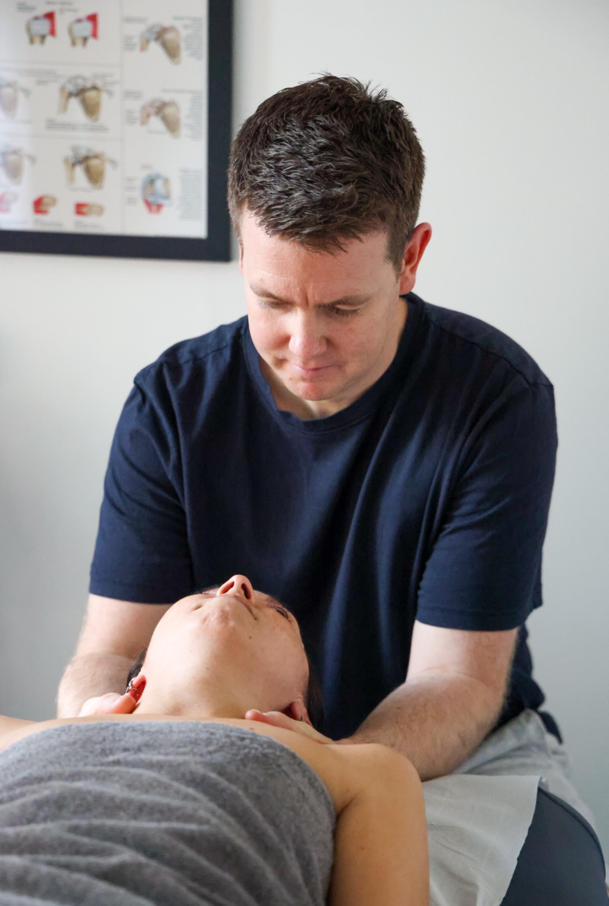 A man providing a neck massage to a young person lying on a treatment table, with a medical poster visible on the wall in the background.