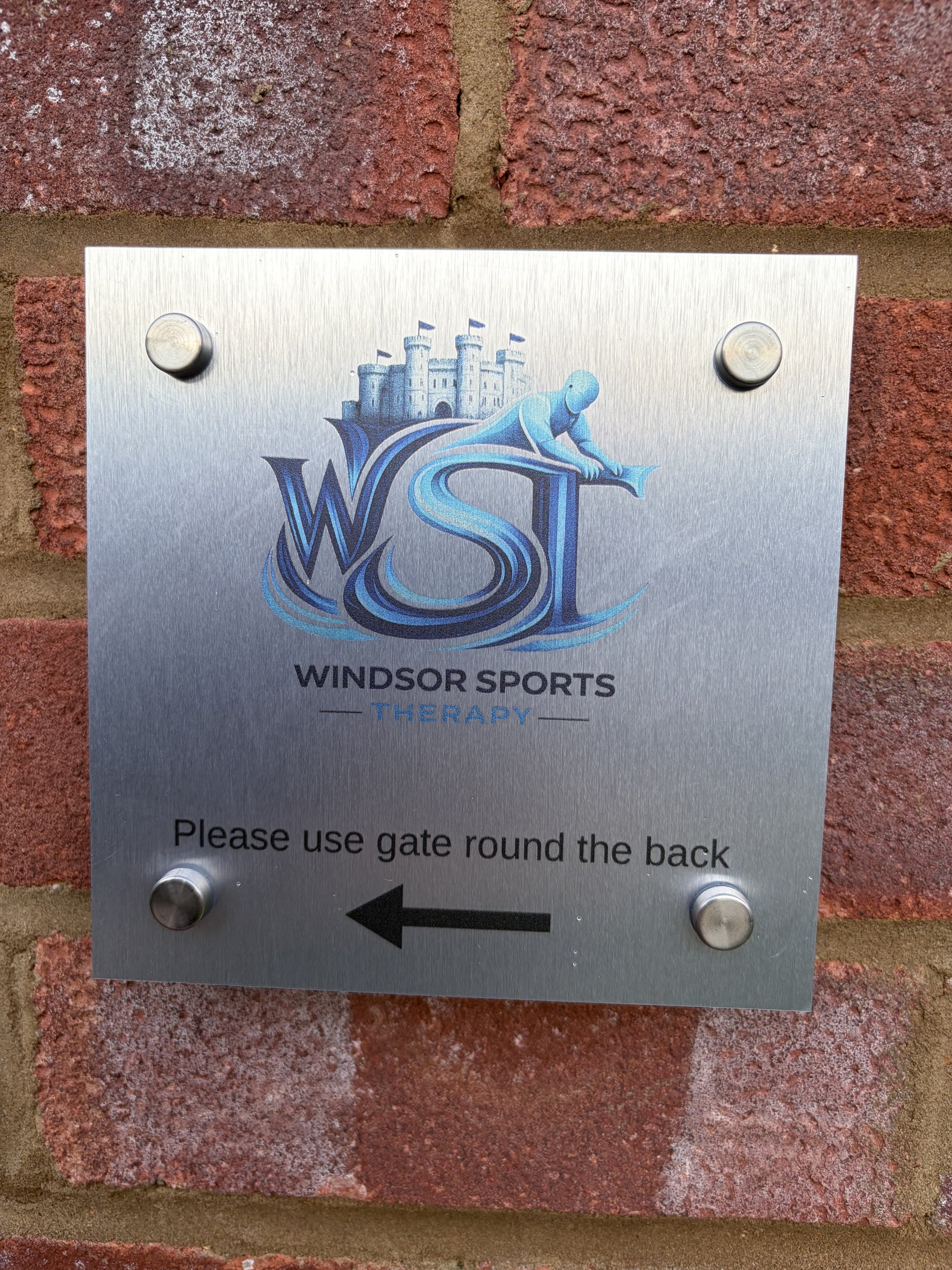 A metal sign attached to a brick wall for Windsor Sports Therapy, with a logo of a castle, and an arrow pointing left with the text 'Please use gate round the back.'