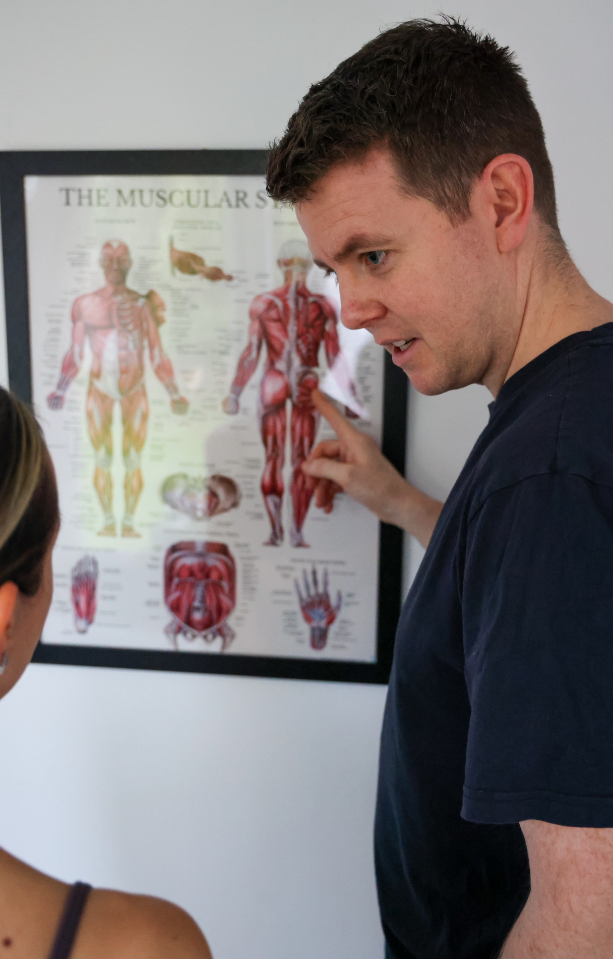 Man pointing at a muscular anatomy poster on the wall while explaining to a woman.