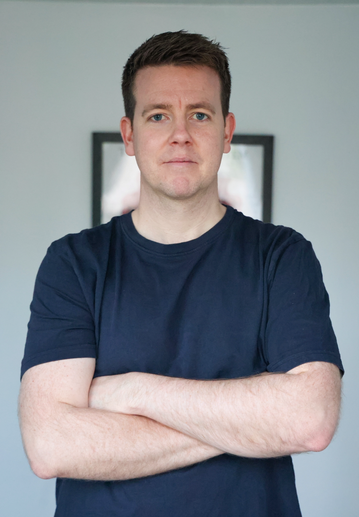 A young man with short dark hair and blue eyes, wearing a dark blue t-shirt, standing with arms crossed in front of a gray wall with a blurred picture frame behind him.