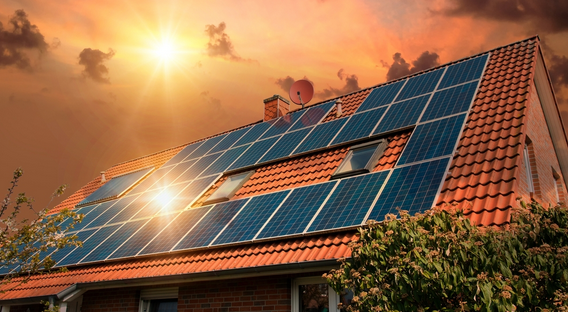 House with solar panels on red-tiled roof during sunset, with some trees in the foreground and a satellite dish on the roof.