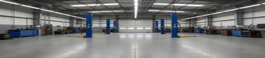An empty industrial workshop or garage with multiple car lifts, workbenches, and tools along the walls, illuminated by fluorescent lighting.