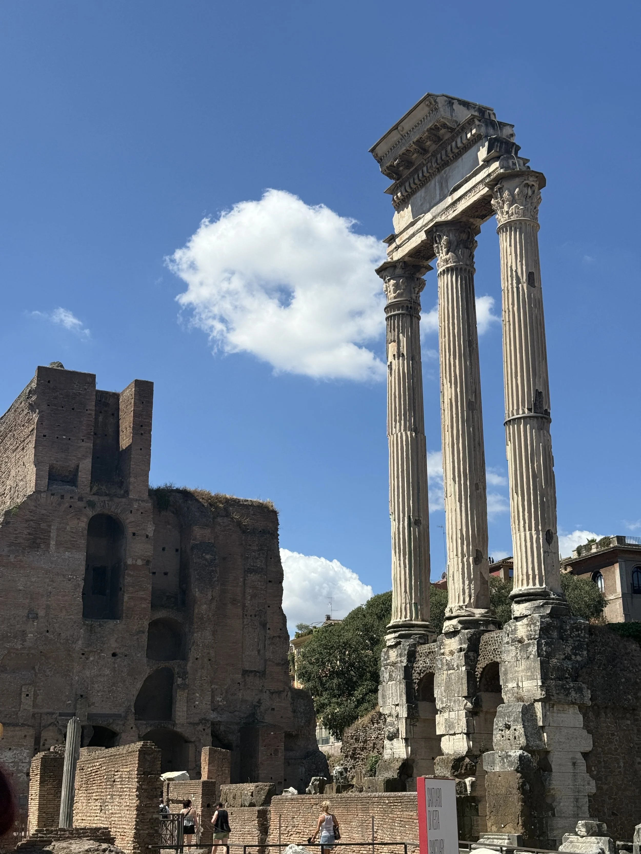 Ancient Roman ruins with three tall, Corinthian columns standing among brick structures and ruins. Blue sky with a few clouds in the background.