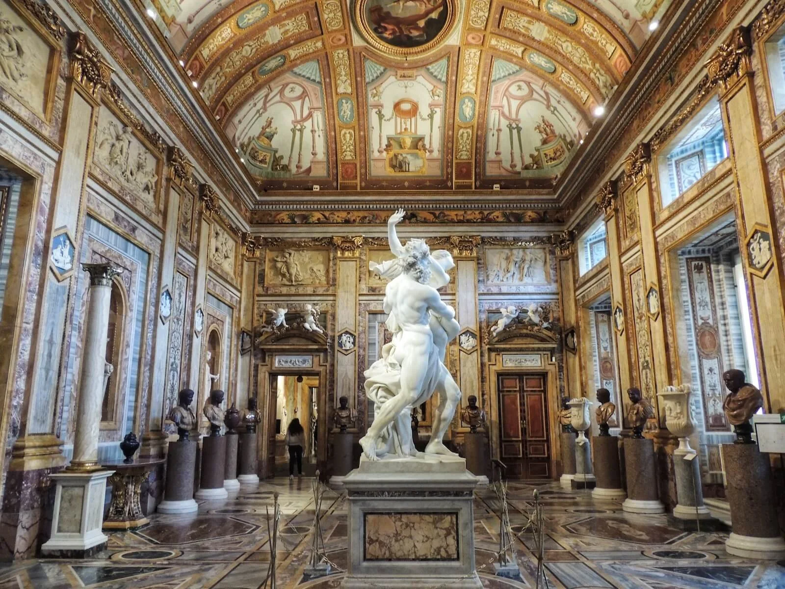 Interior of a lavish museum or palace hall with ornate decorated walls and ceiling, marble bust sculptures on pedestals lining the walls, and a central white marble statue of a mythological figure surrounded by a chain barrier.