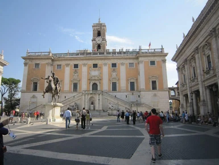 A historic building with stairs and sculptures in a European city square, tourists walking around.