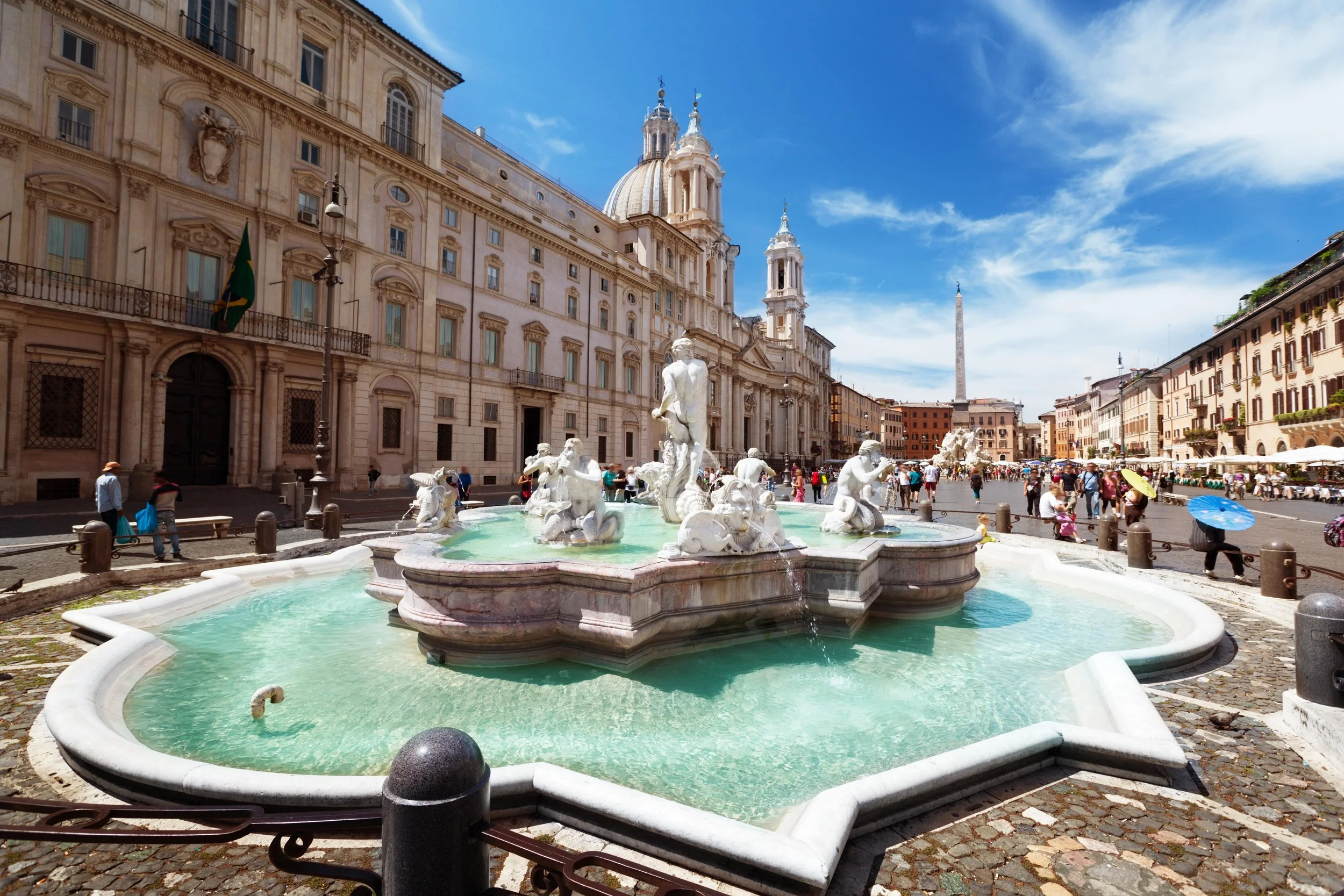 A historic European city square with a decorative fountain featuring statues, surrounded by baroque architecture, with a clear blue sky and a tall obelisk in the background, and people walking with umbrellas.