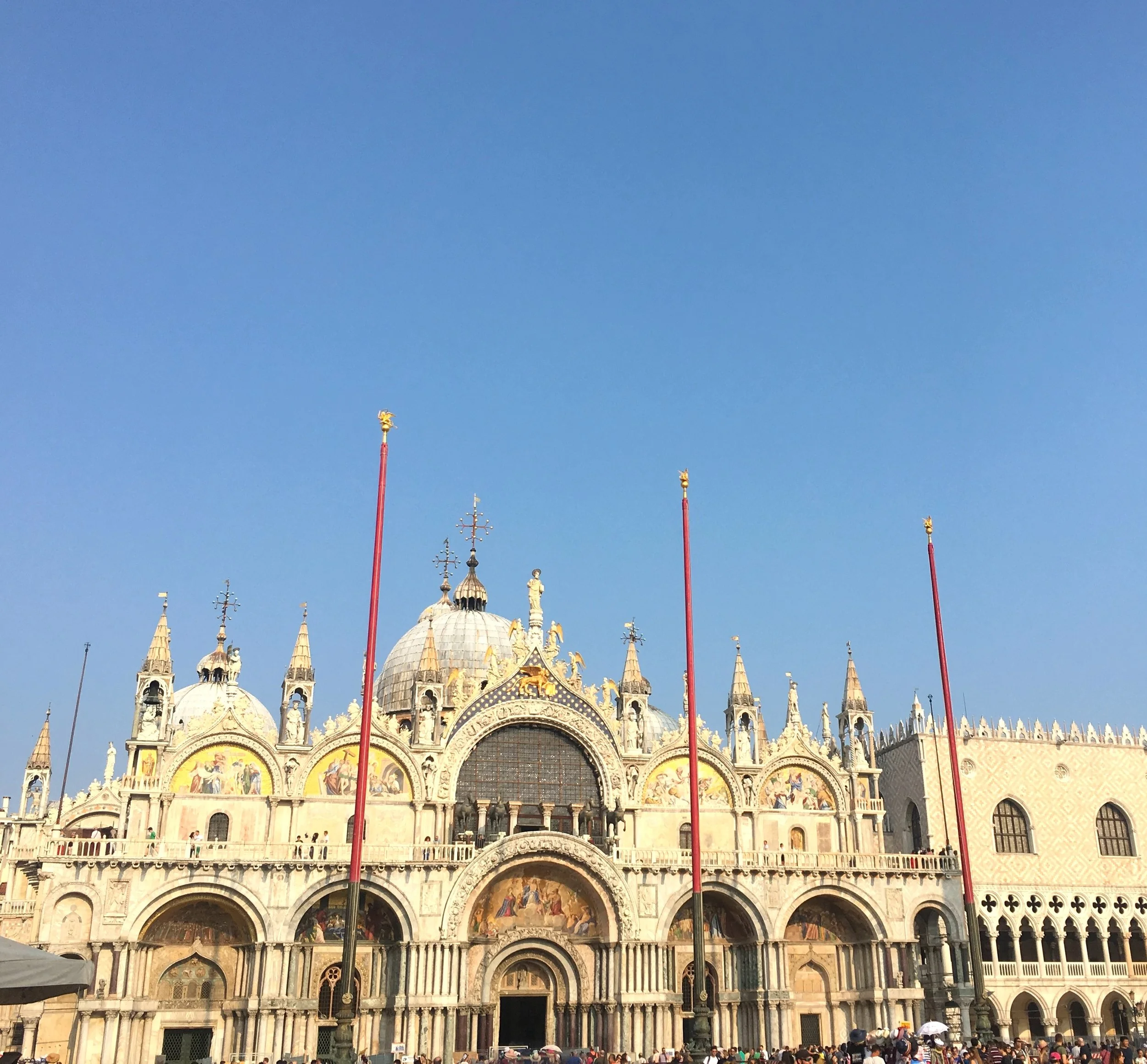 View of St. Mark's Basilica in Venice, Italy, with clear blue sky, flags, and people gathered in front.