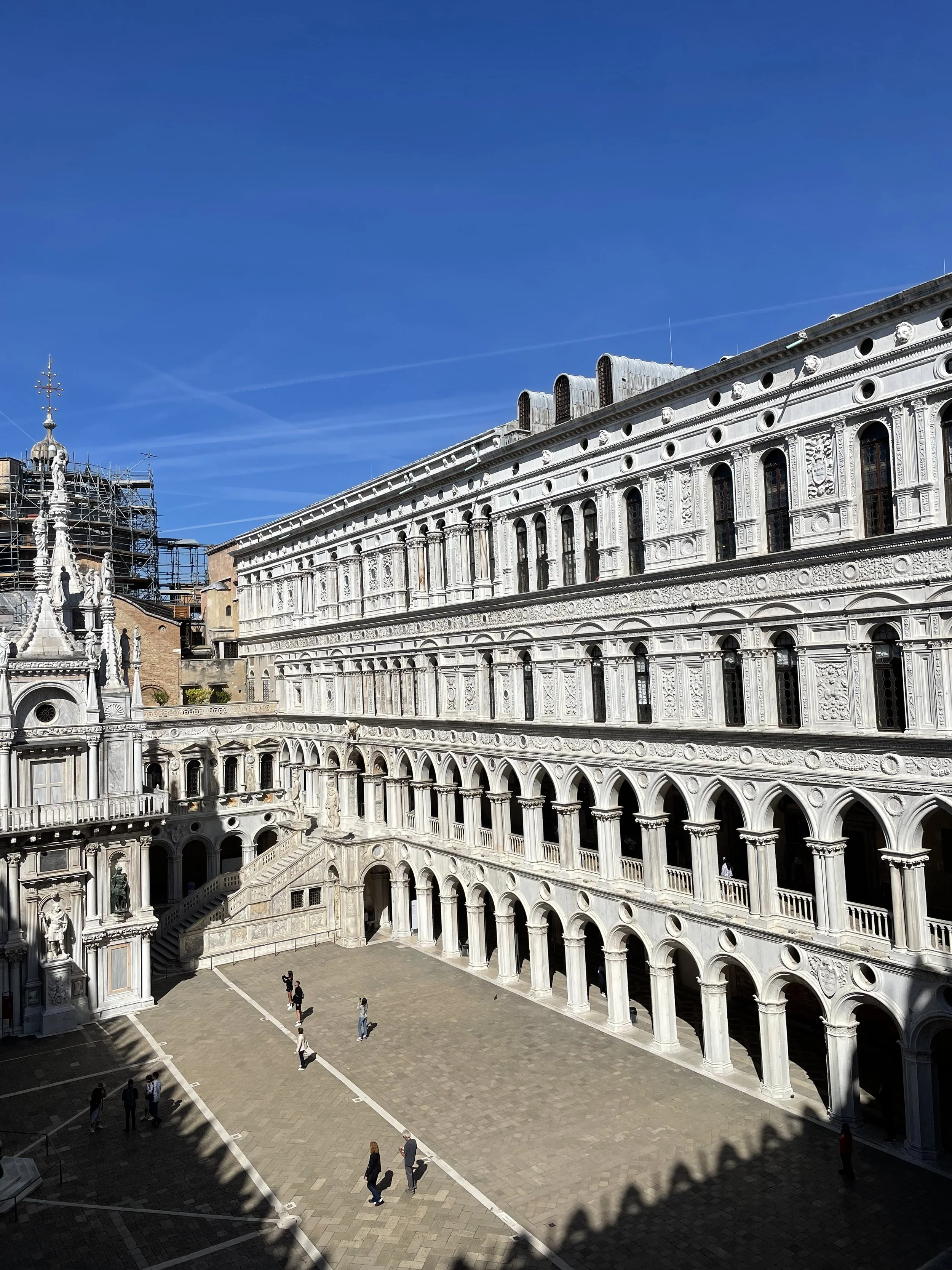 A historical building with ornate white stone architecture and arched windows, with people walking in the courtyard beneath a clear blue sky.