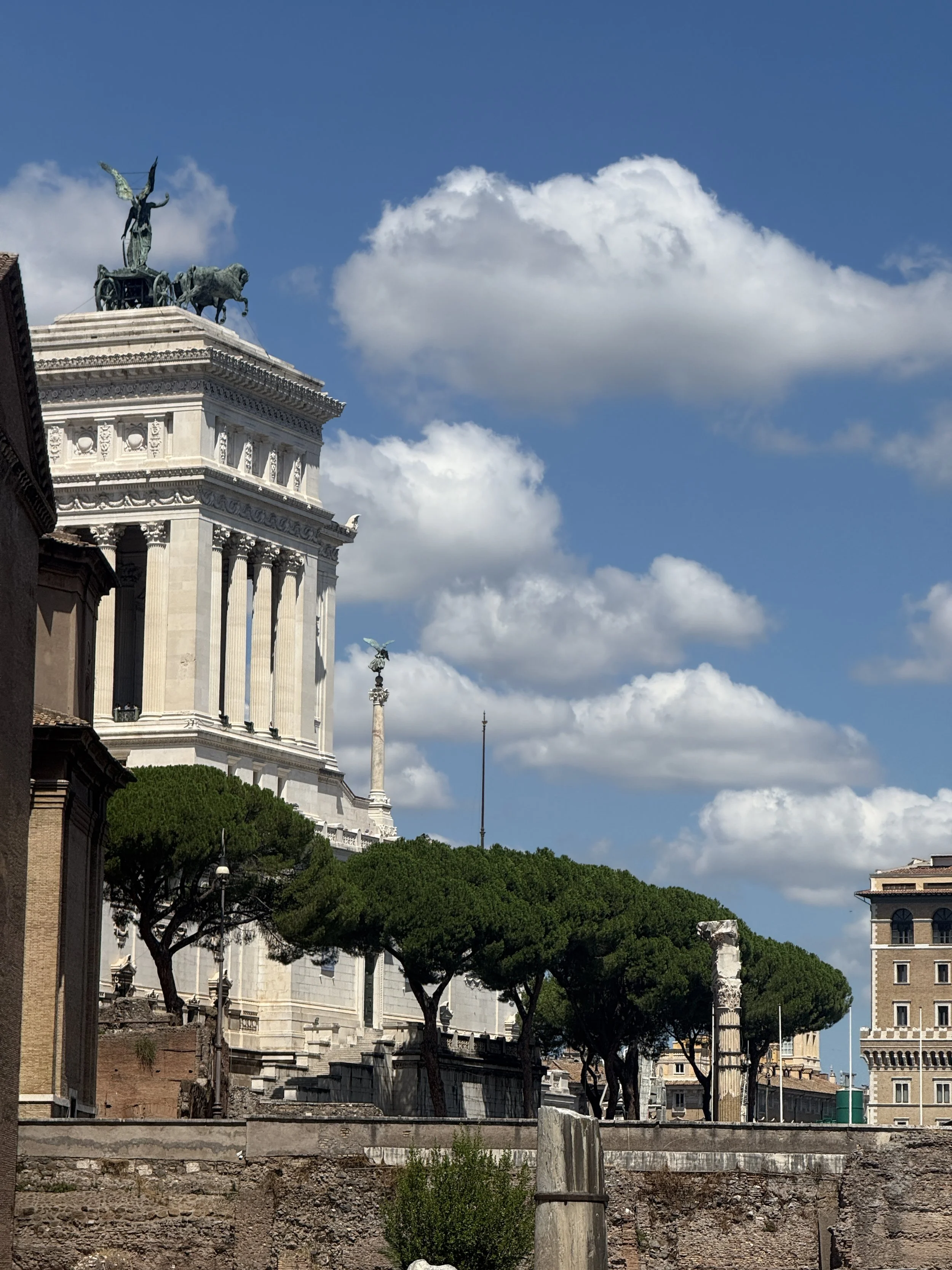 A historic white marble monument with a statue on top of a chariot pulled by horses, set against a bright blue sky with scattered clouds and green trees in the foreground.