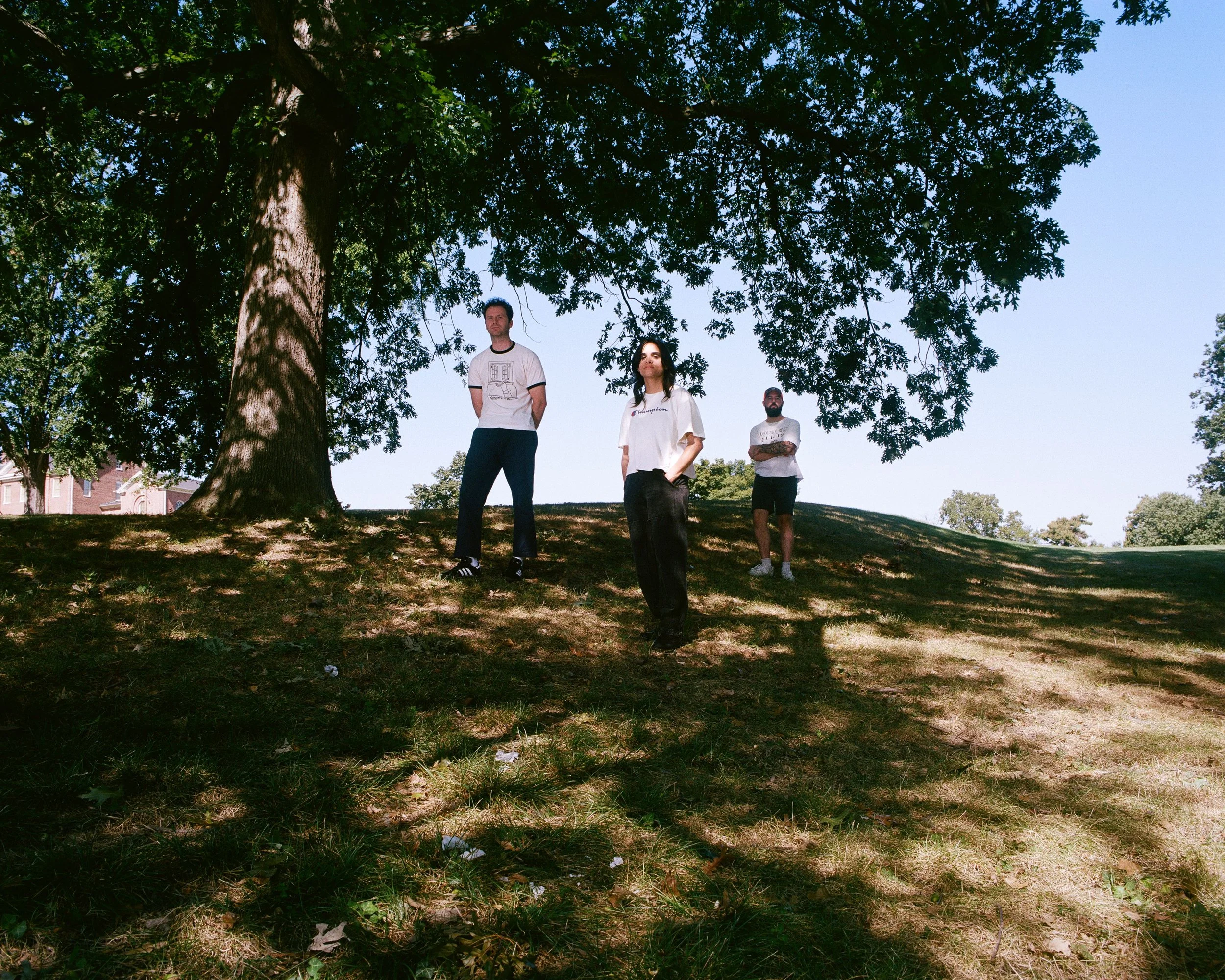 Three individuals standing under a large tree in a park, with clear blue sky overhead.