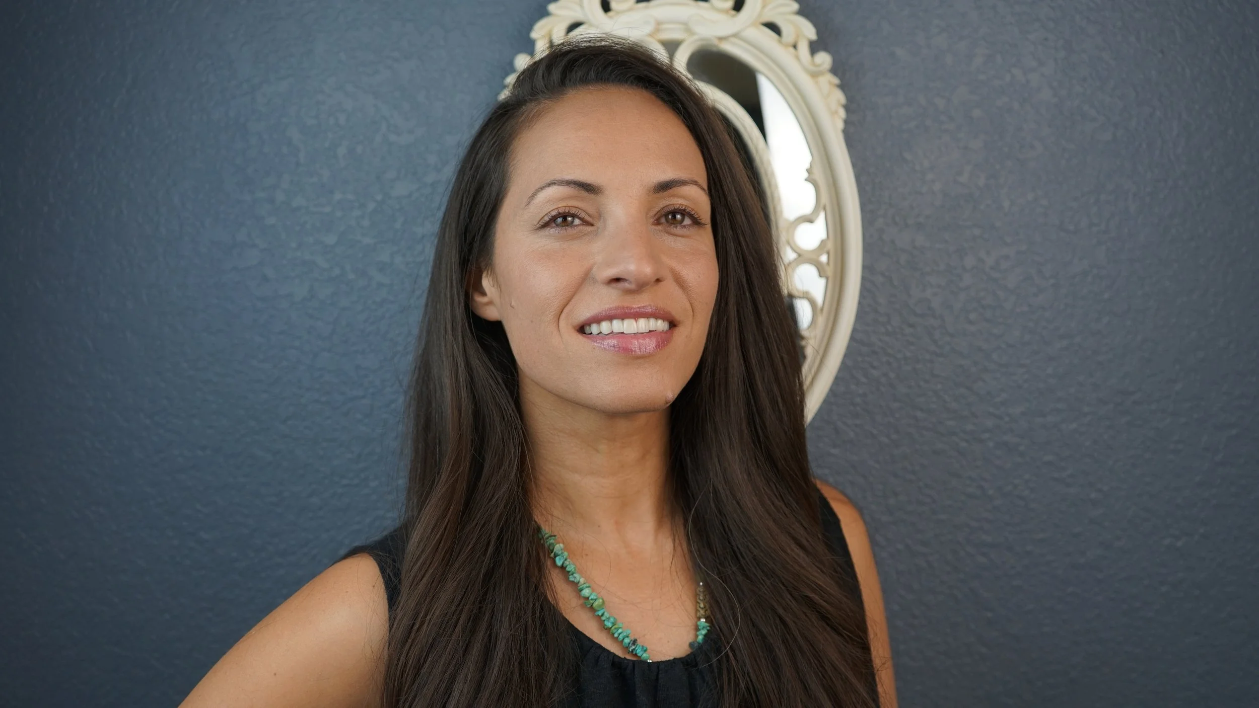 A woman with long dark brown hair, smiling, wearing a black sleeveless top and a turquoise beaded necklace, standing in front of a dark gray wall with a decorative white mirror in the background.