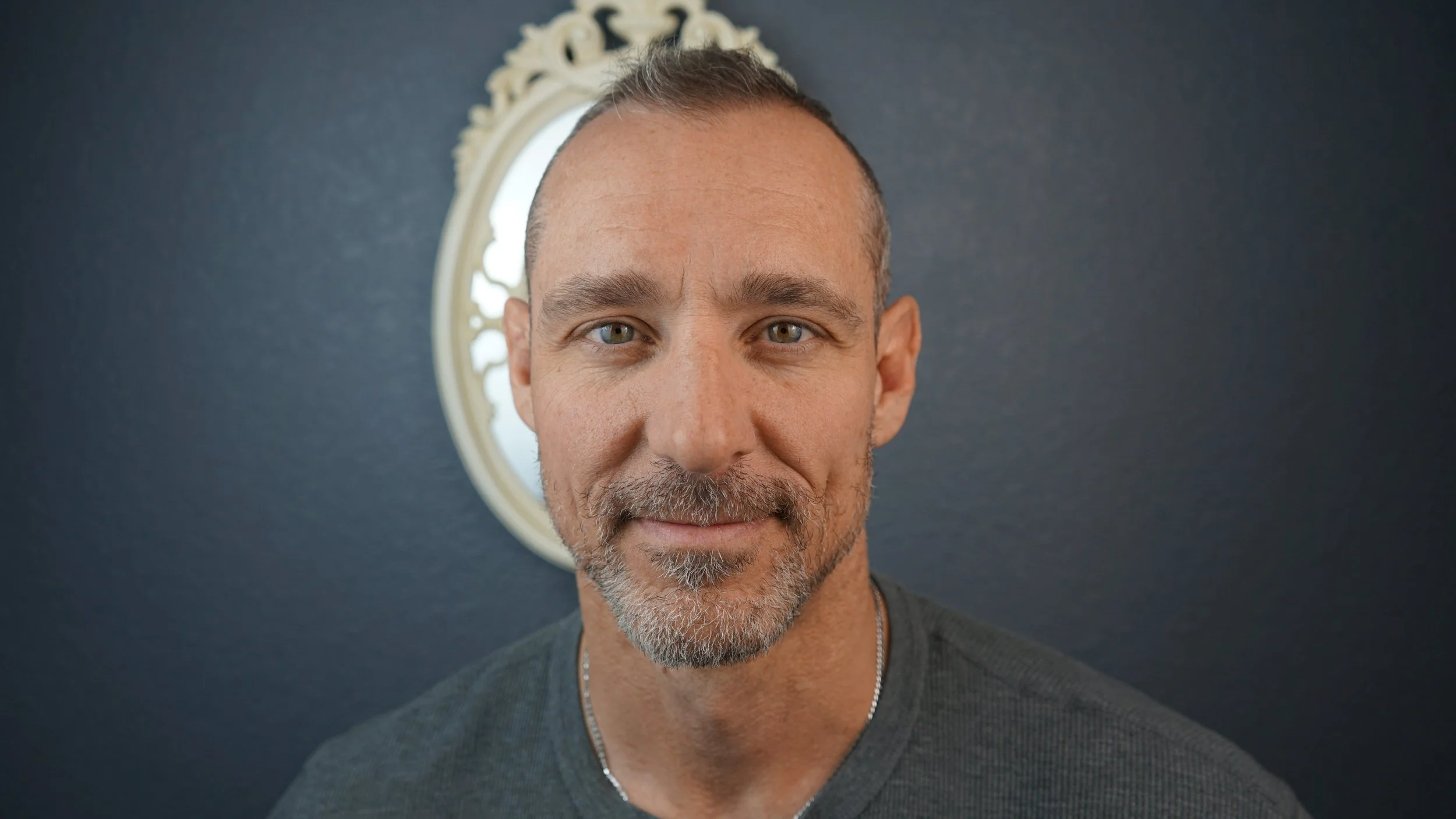 Close-up portrait of a middle-aged man with short hair and a beard, looking at the camera with a slight smile, standing in front of a dark wall with a mirror