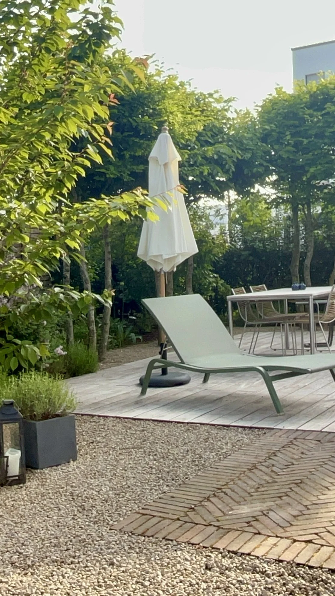 Ein Garten mit Liegestuhl, Sonnenschirm, Esstisch und Stühlen, umgeben von Bäumen und Pflanzen, sonnig.