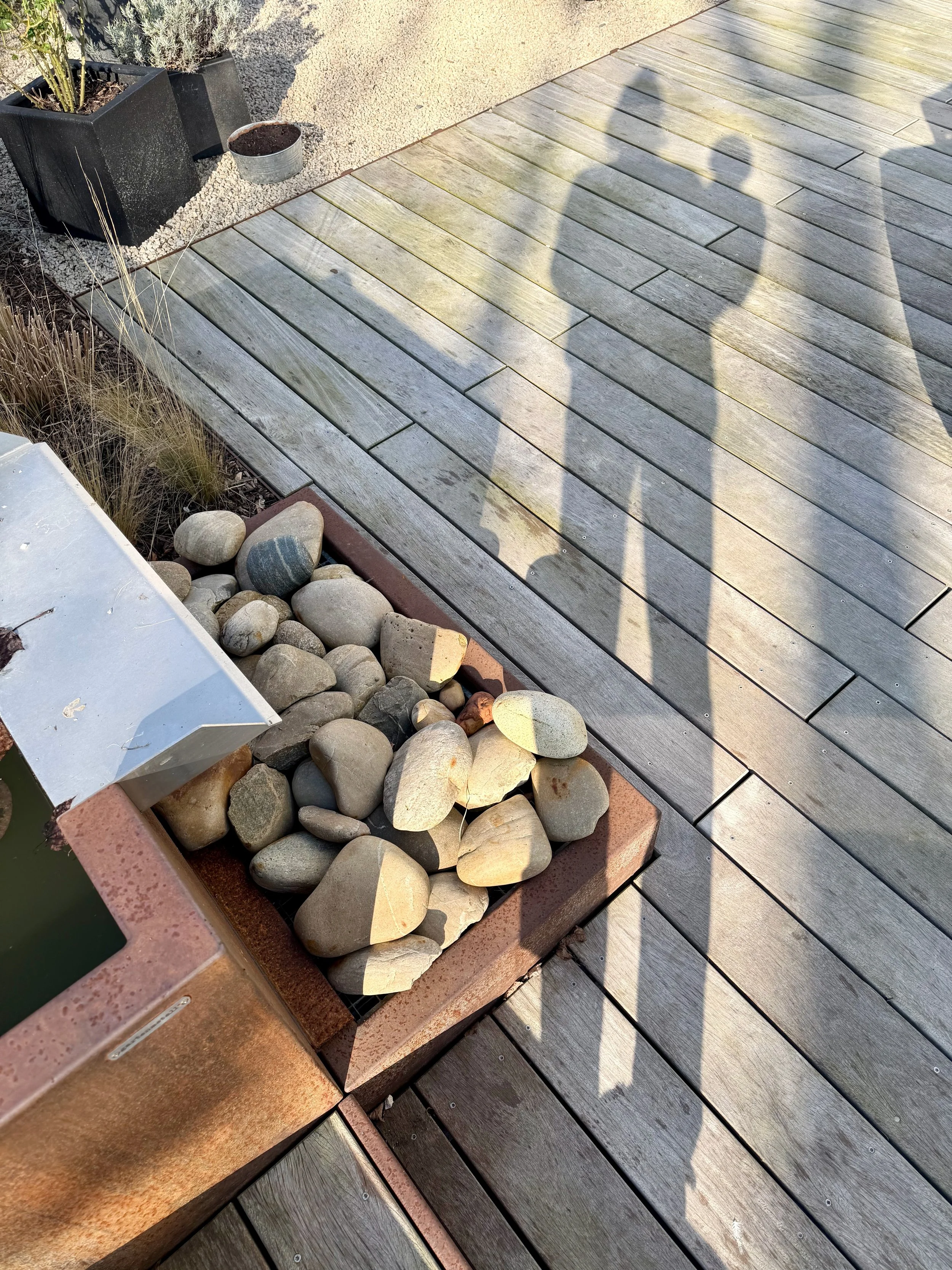 Ein Garten mit Holzdielenboden, einem Pflanzkübel mit Steinen, einem kleinen Wasserbecken und Staudenpflanzen. Schatten von drei Menschen auf dem Boden.