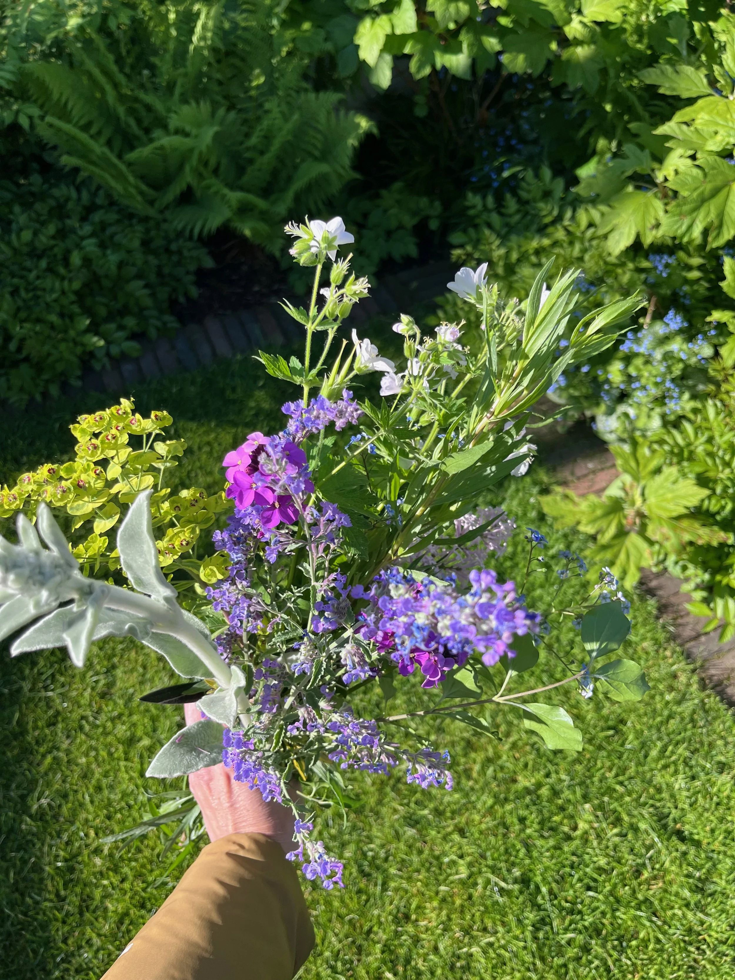 Person hält einen Blumenstrauß mit weißen, violetten, lilafarbenen und grau-grünen Blumen in einer Hand, mit grünem Rasen im Hintergrund