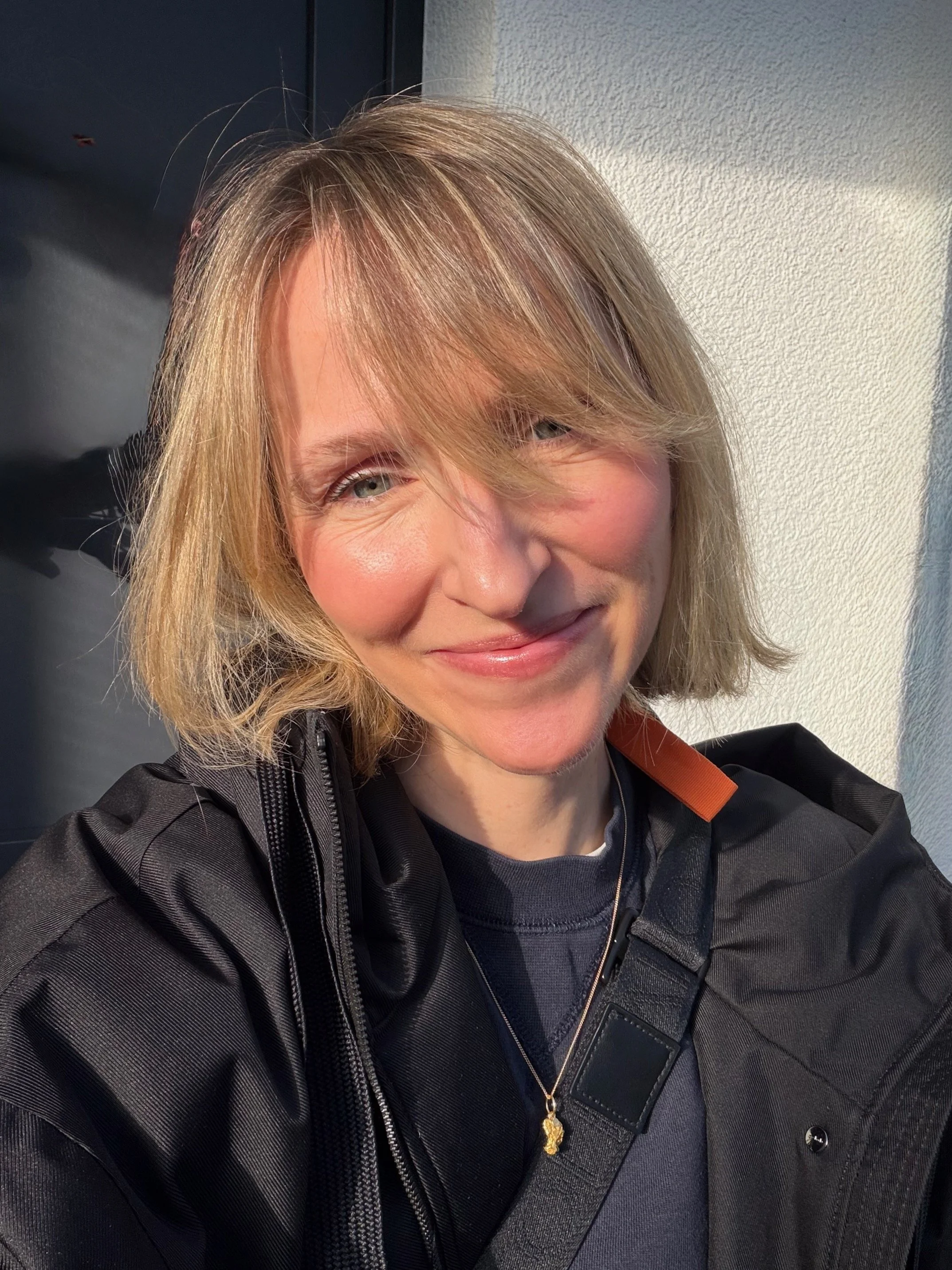 Nahaufnahme eines lachenden Frauenkopfes mit blonden, gestreiften Haaren, die teilweise das Gesicht bedecken, vor einem weißen Wand und schwarzem Türrahmen, getragenem schwarzen Jacke, goldener Halskette und Smartphone im schwarzen Halfter