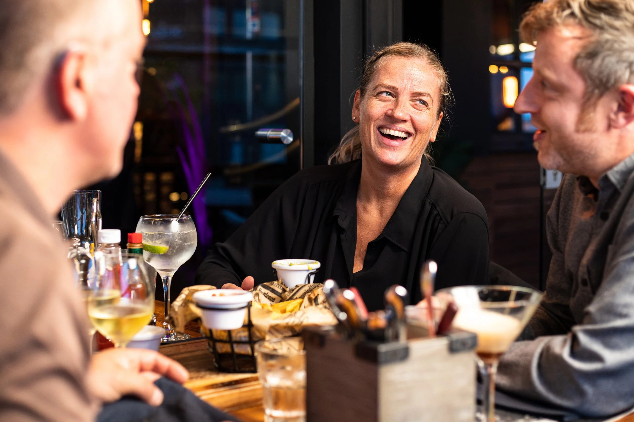 Fire mennesker som smiler og snakker på en restaurant eller bar, med drinker og mat på bordet.