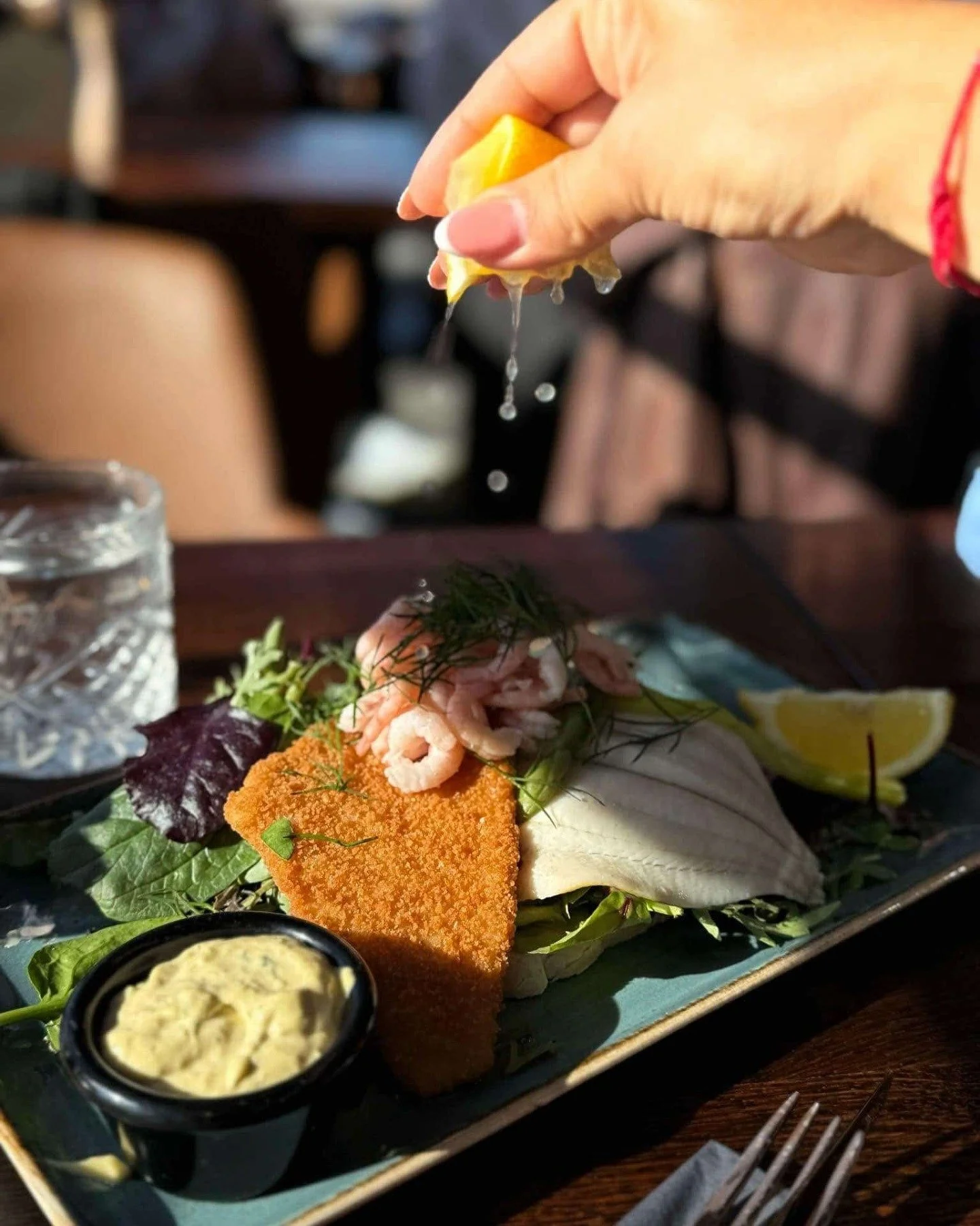 Person som holder en sitronskive over en tallerken med rekesalater, fisk og grønnsaker, med en liten skål med aioli.