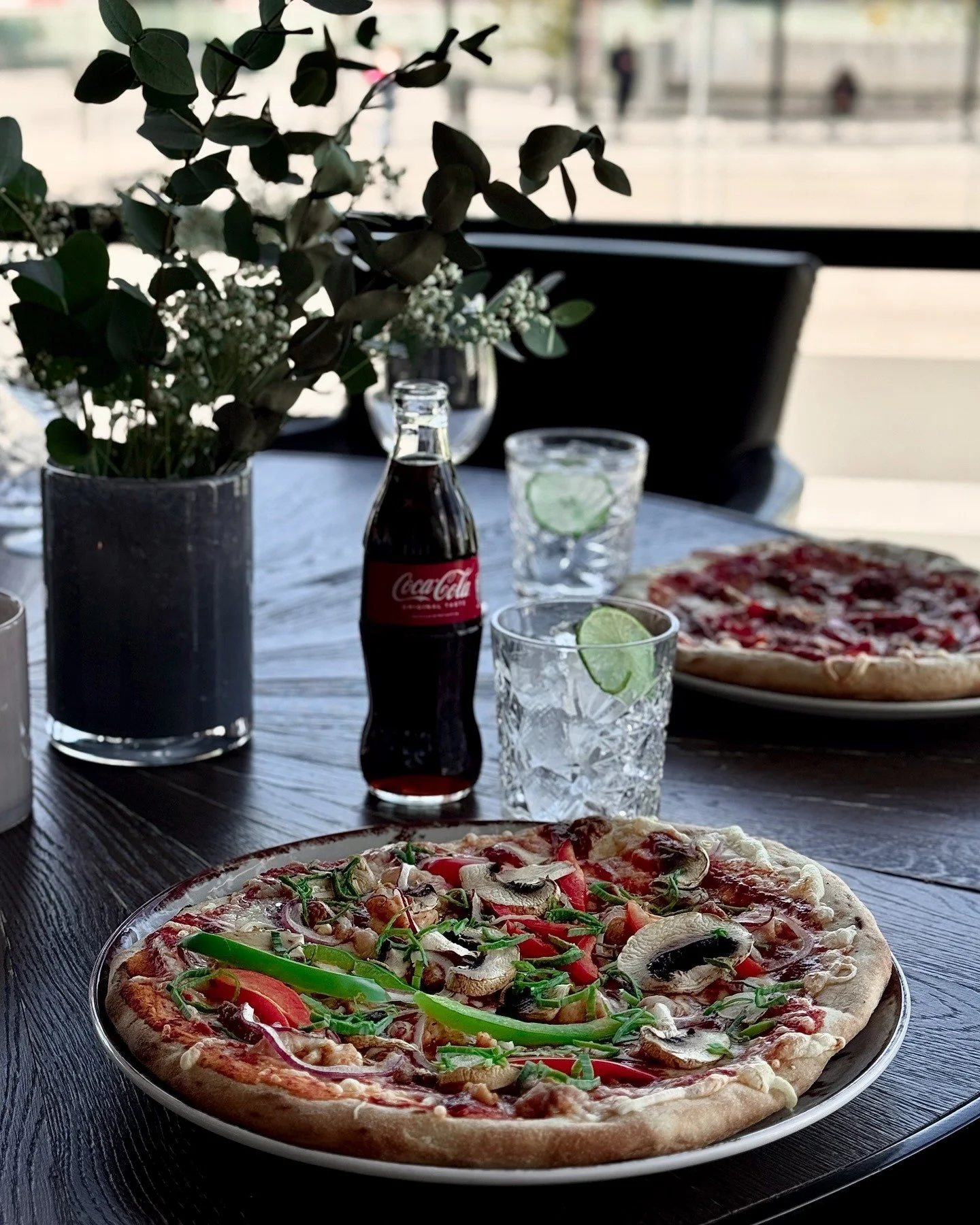 To pizzaer på en restaurantbord, med en Coca-Cola-flaske, to glass med limebiter, og en stor blomsterdekorasjon.