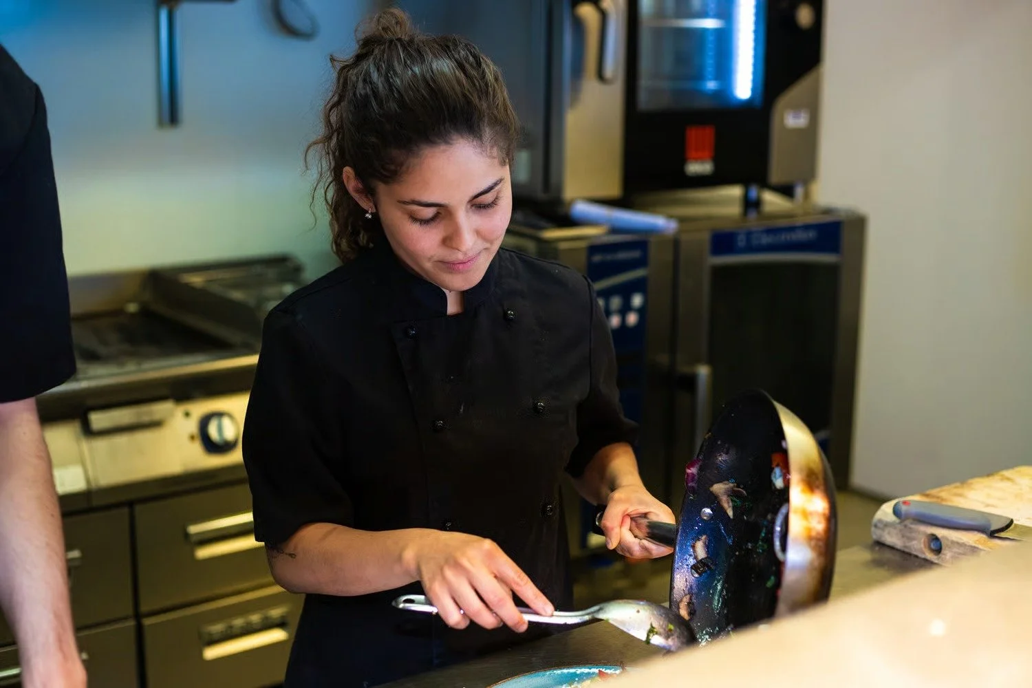 En kvinne i svart kokketøy som lager mat på en restaurant kjøkken. Hun bruker en stekepanne og et skjærebrett ligger på benken.