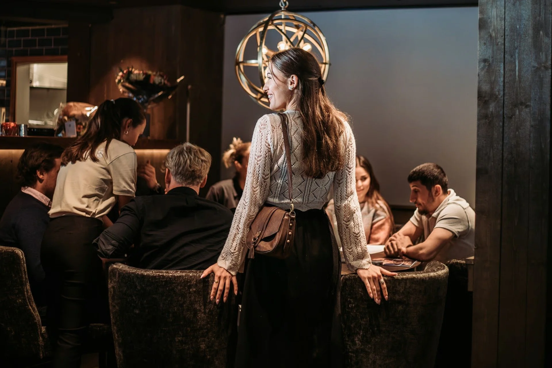 En kvinne står bak en gruppe mennesker som sitter rundt et bord i en restaurant eller kafé. Det er en mørk og koselig innredning med en dekorativ lysekrone i taket.