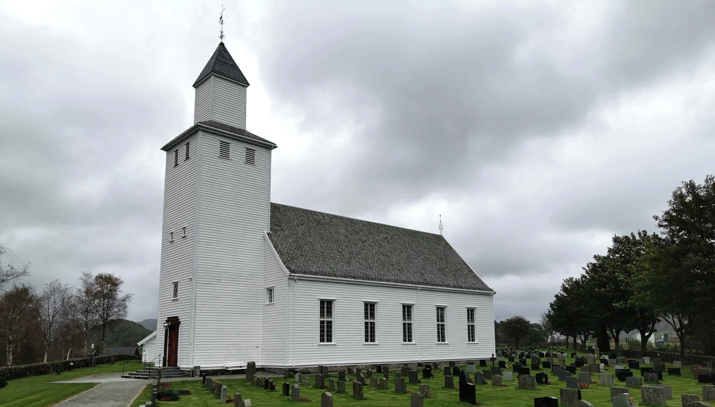 Oversiktsbilde av en hvit kirke med kirkegård foran, omgitt av trær og overskyet himmel.