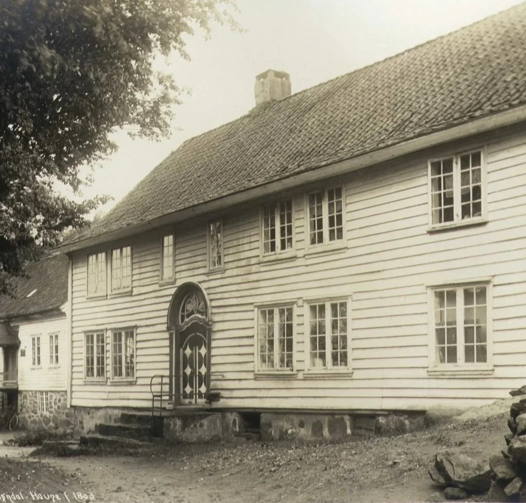 En historisk trehus med hvitmalte vegger og flere vinduer, antagelig fra 1800-tallet, med en steinmur og trapp ved inngangsdøren.
