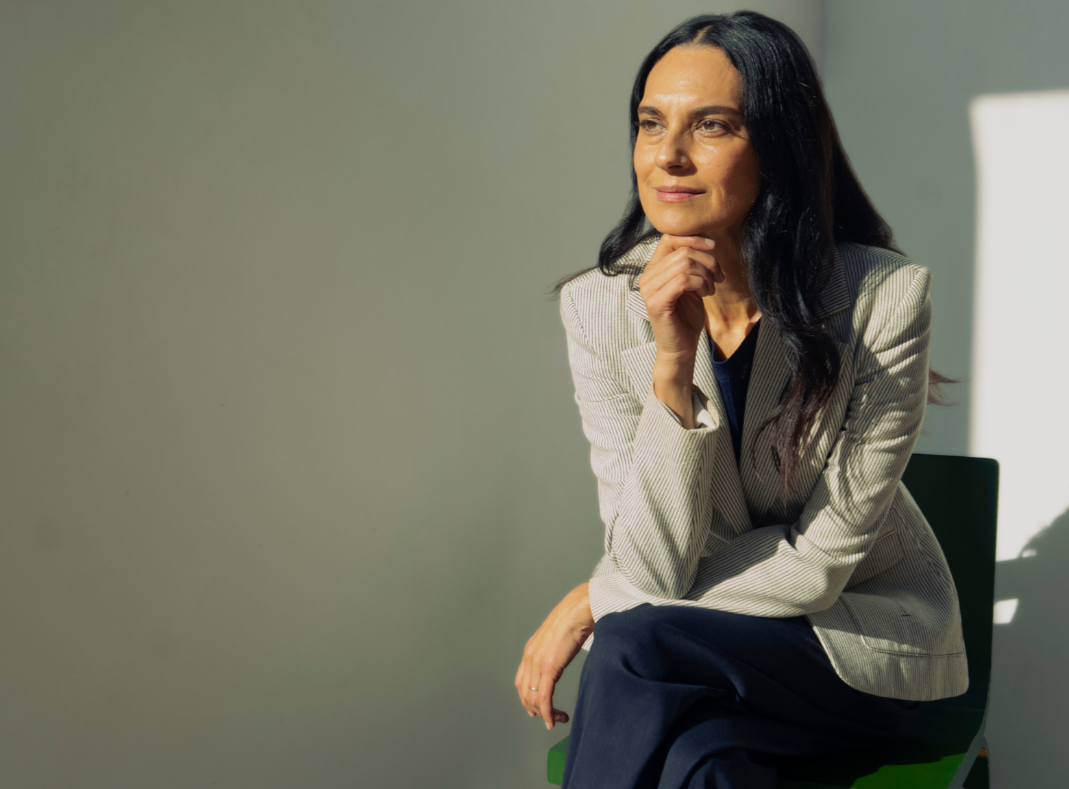Sarvia Jasso Art Advisor sitting on a green chair, wearing a light-colored blazer and dark pants, thinking with her hand on her chin. Admiring the beauty of art in all of its differences.