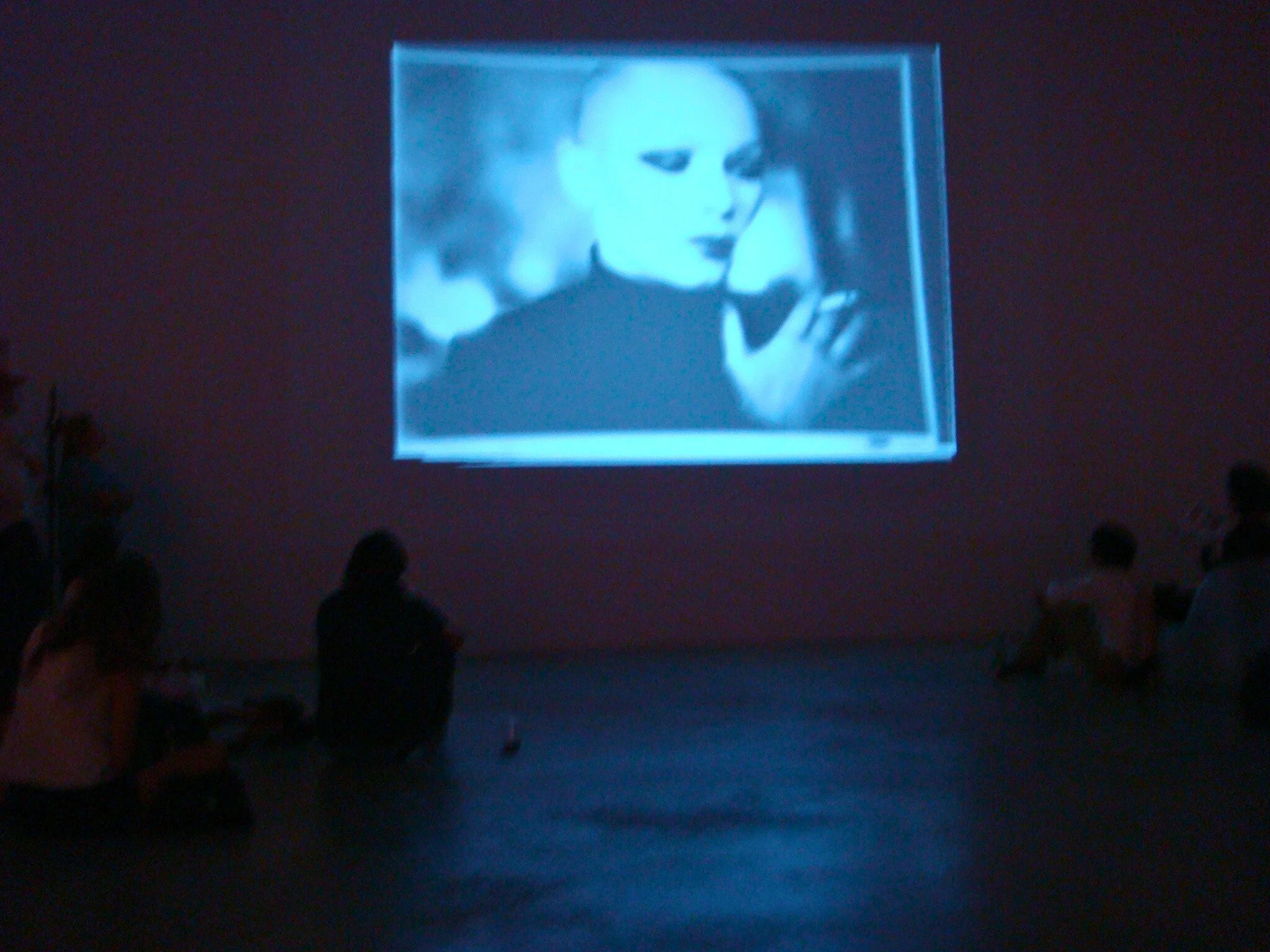 People sitting on the floor watching a black-and-white movie or video projected on a wall for screening of Manon and androgyny.