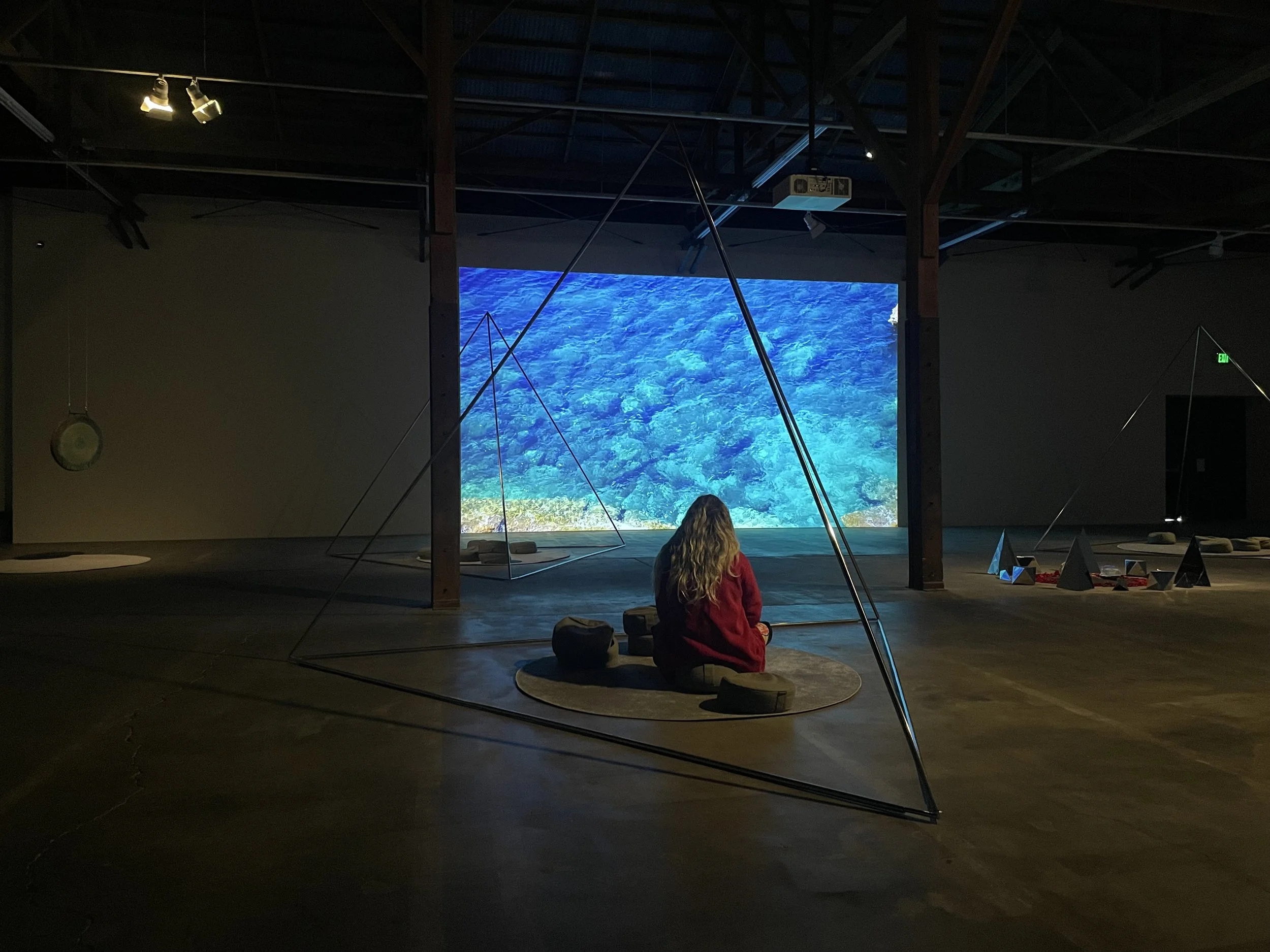A woman with long blonde hair in a red jacket sitting cross-legged on a circular rug and meditating in front of a large digital screen displaying an underwater scene of blue ocean water by artist Kathryn