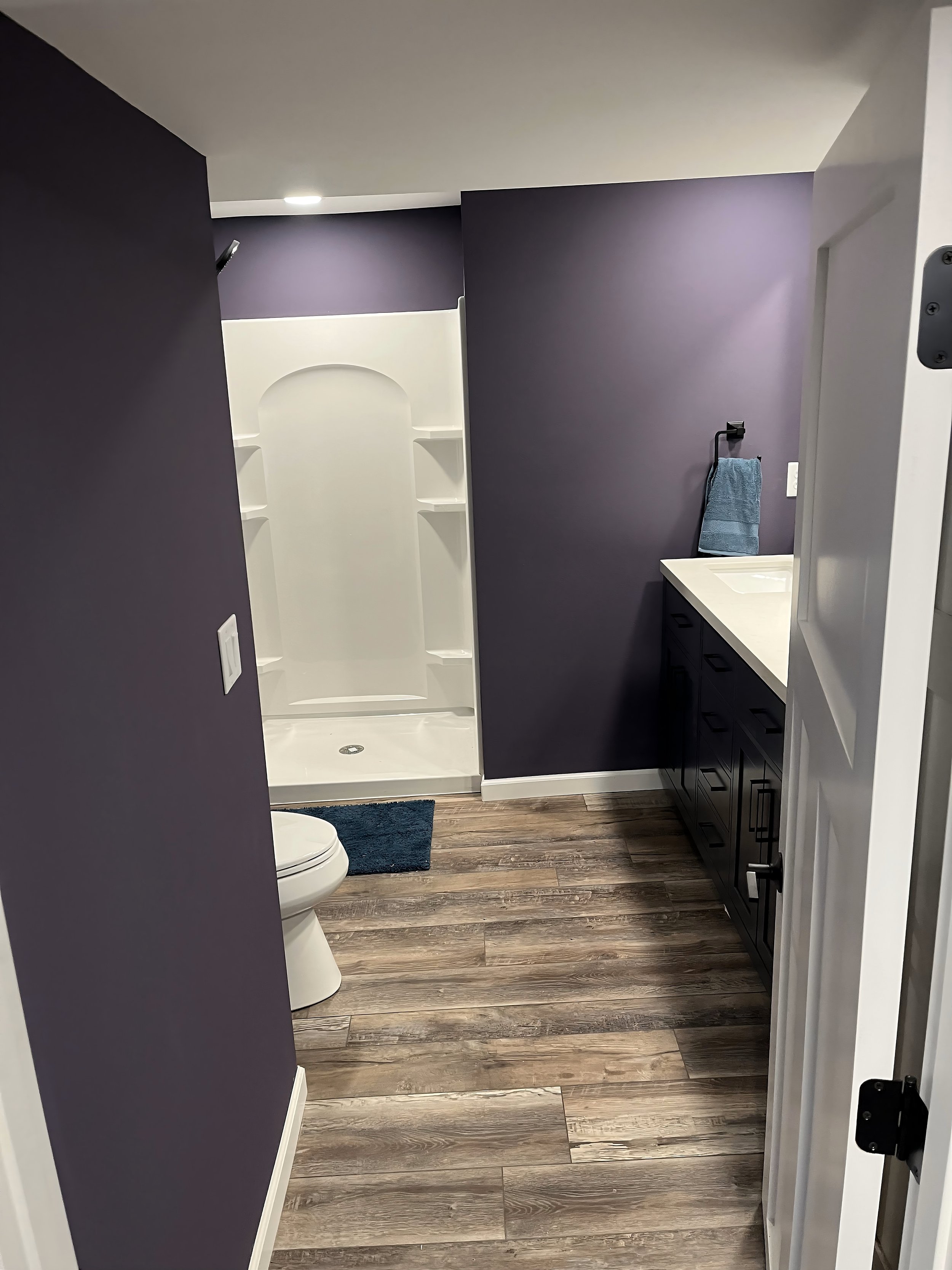 A modern bathroom with purple walls, a white shower with built-in shelves, a toilet, a dark blue rug, a dark vanity with a white countertop, and a blue towel hanging on a black towel holder.