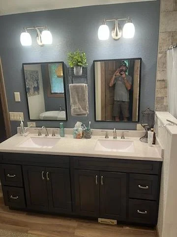 Bathroom vanity with double sinks, dark cabinet, and white countertop. Two mirrors with light fixtures above and a small plant on the wall between them. A person taking a photo is visible in the reflection.