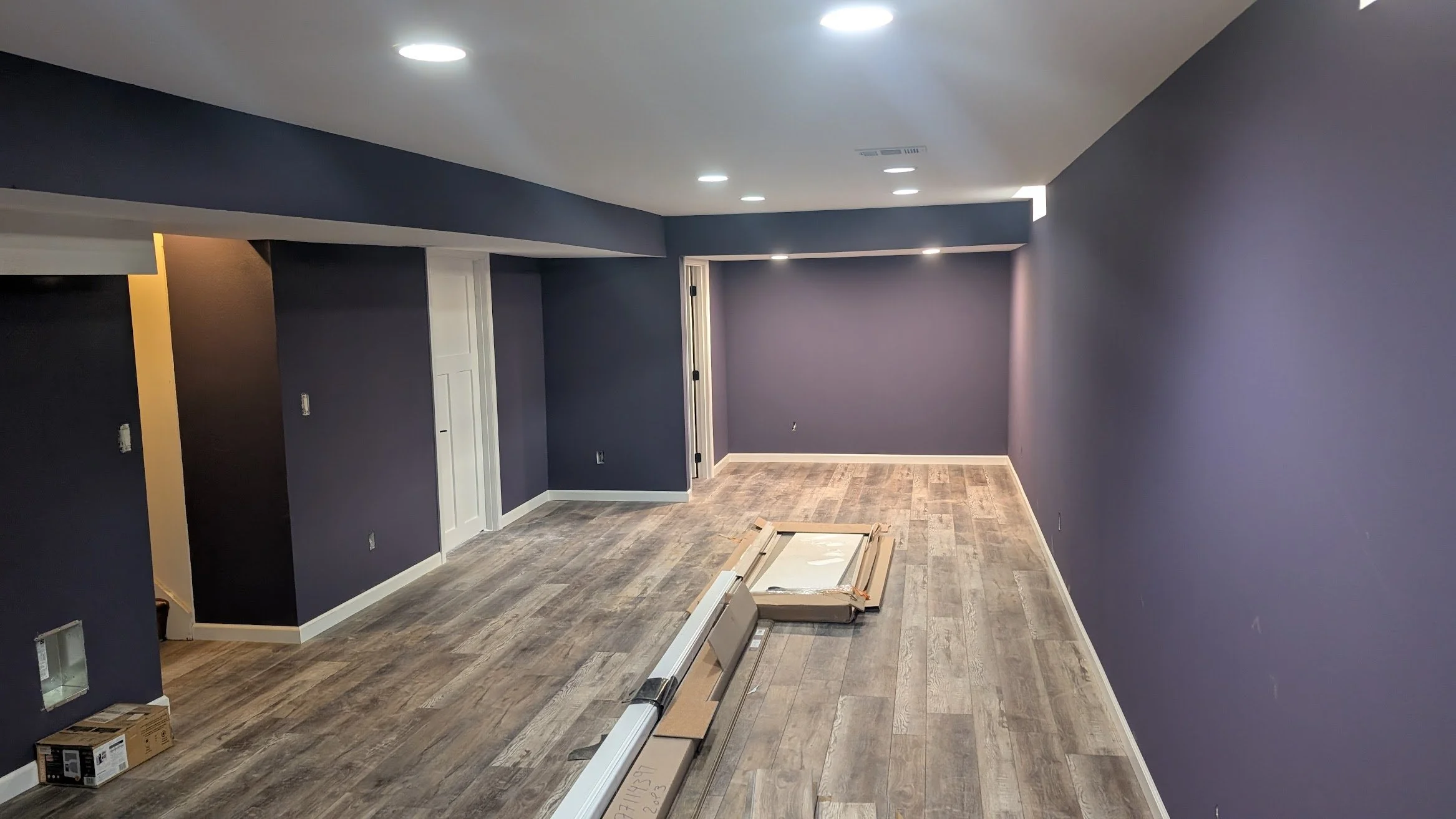 Empty room with dark purple walls, wood-look flooring, recessed ceiling lights, some uninstalled baseboards, and unopened boxes on the floor.