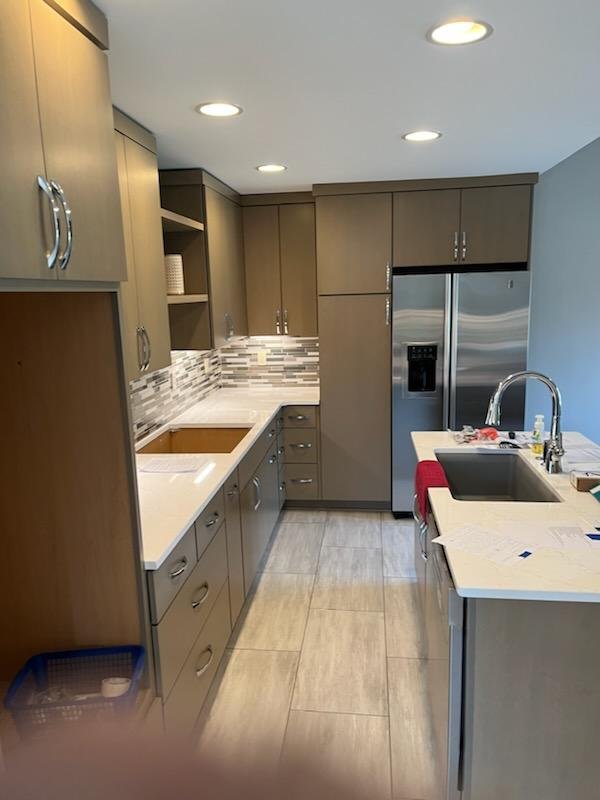 Modern kitchen with gray cabinetry, stainless steel refrigerator, white countertops, mosaic tile backsplash, and a black sink with a faucet.