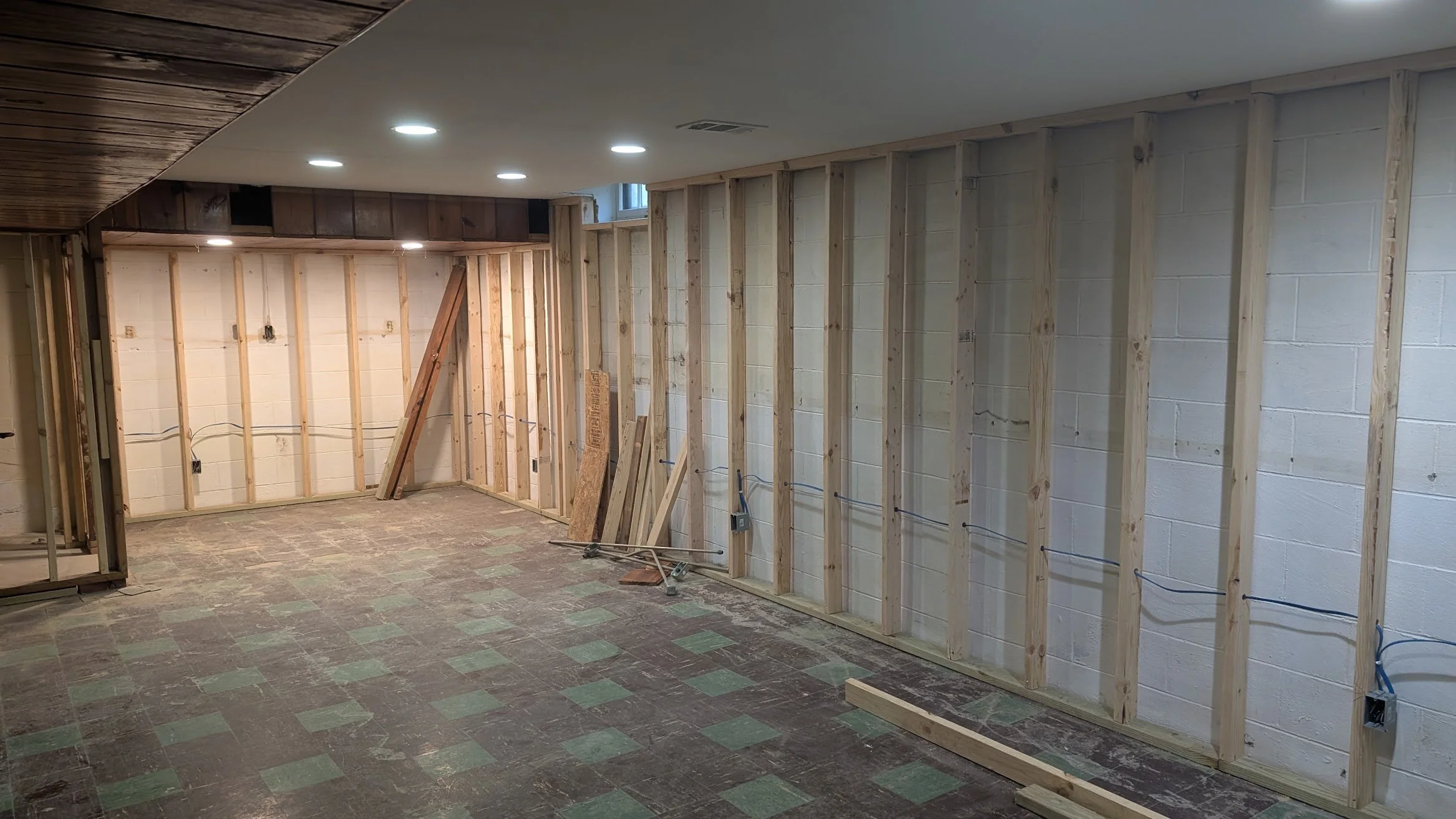 Interior of a basement under renovation with exposed wooden wall studs, electrical wiring, and partially finished ceiling with recessed lighting.