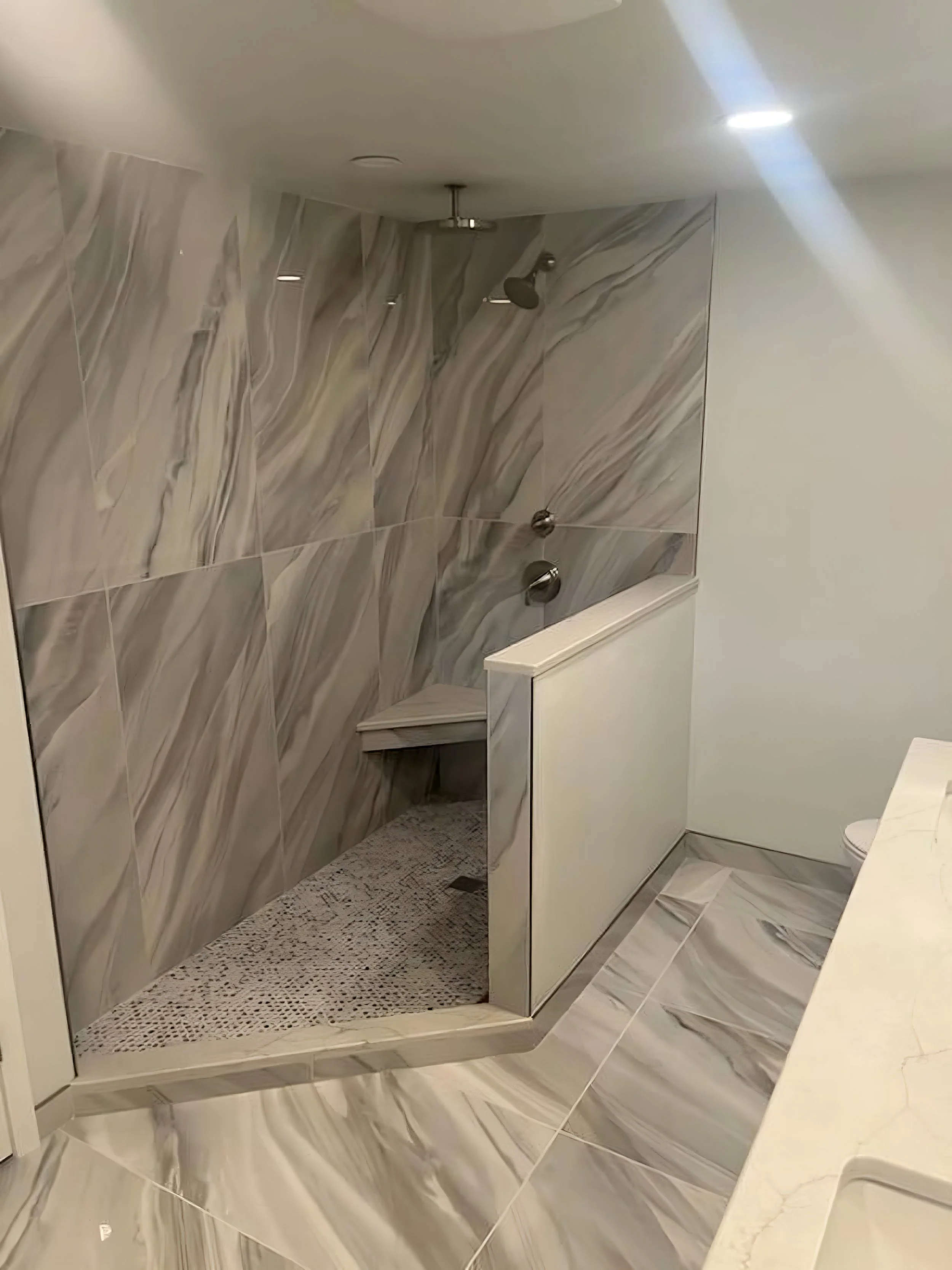 Marble-tiled shower with two showerheads, a built-in bench, and a partition wall with marble tile on the floor and walls.