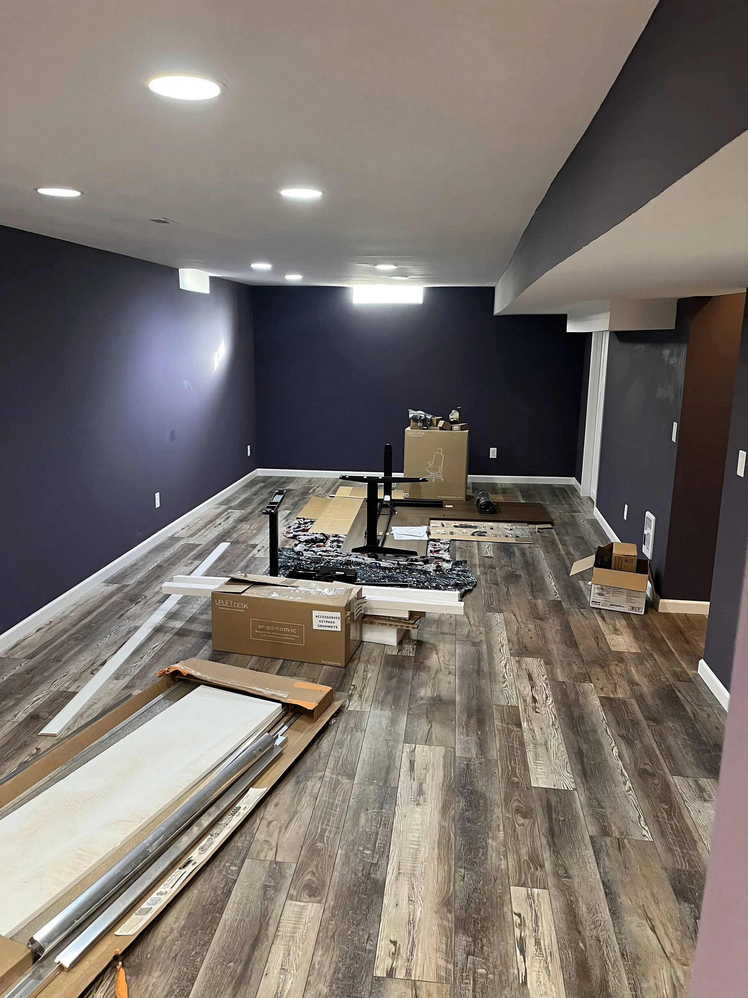 A room under construction or renovation with dark purple walls, wooden flooring, scattered boxes, and tools, including a rolled-up chair base, and construction materials on the floor.