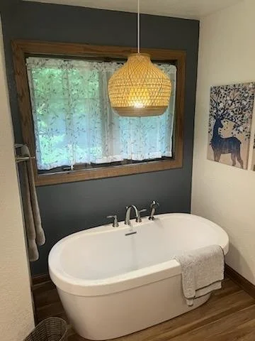 A cozy bathroom with a white freestanding bathtub, a window with lace curtains, a wooden framed window, a hanging light fixture, a towel on the bathtub, and a deer-themed artwork on the wall.