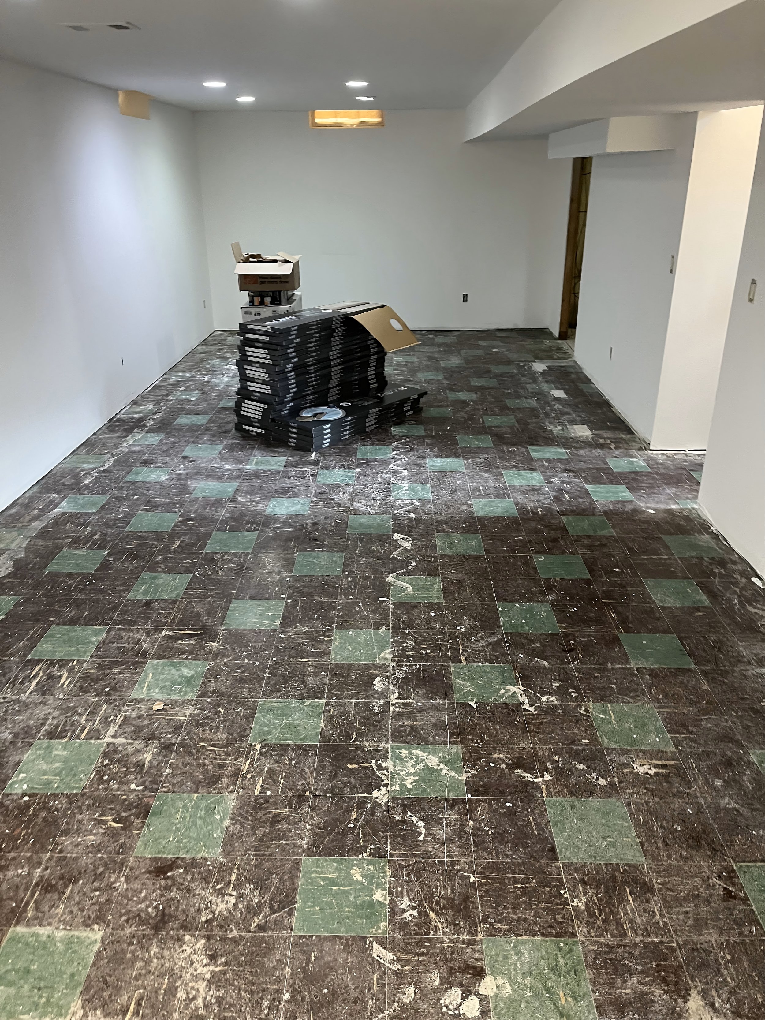 Empty room with white walls, green and brown tiled floor, and ceiling lights. There are stacks of black items and some boxes in the corner.