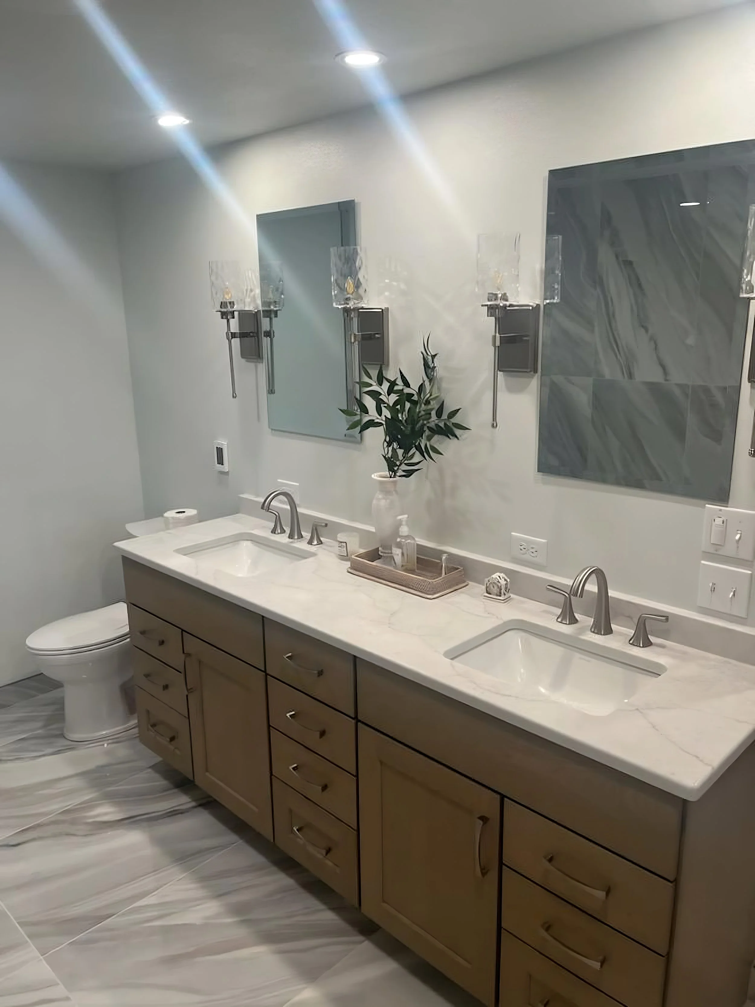 A modern bathroom with a double vanity, two sinks, and two mirrors with wall-mounted lights. The vanity has a beige cabinet with six drawers, a white marble countertop, and a decorative vase with greenery. There is a toilet and marble flooring, and t