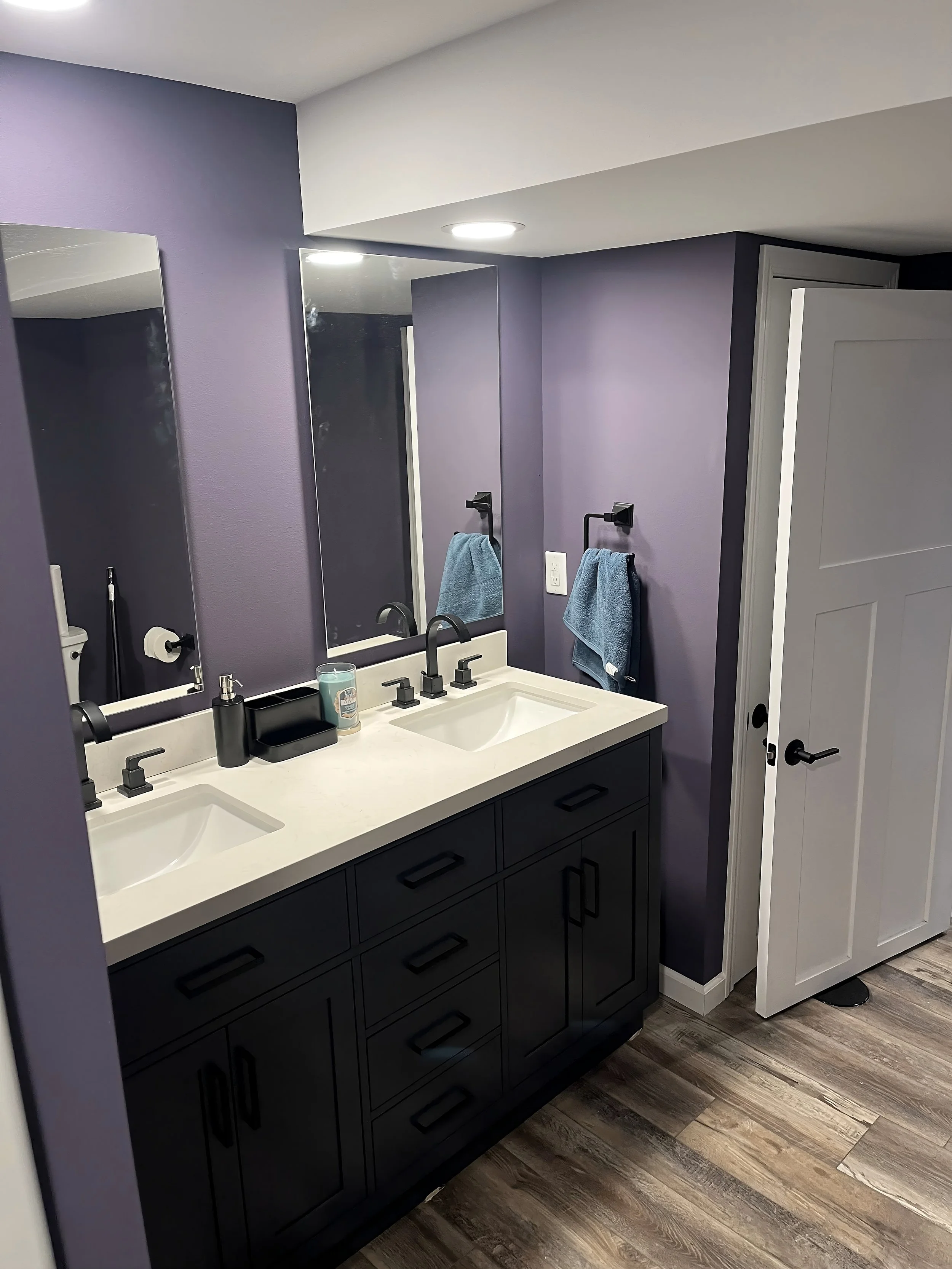 A modern bathroom with a black double vanity, white countertop, and large mirror. Purple walls, blue towels hanging on black towel rings, and a door to the right. Wooden flooring and ceiling lights.