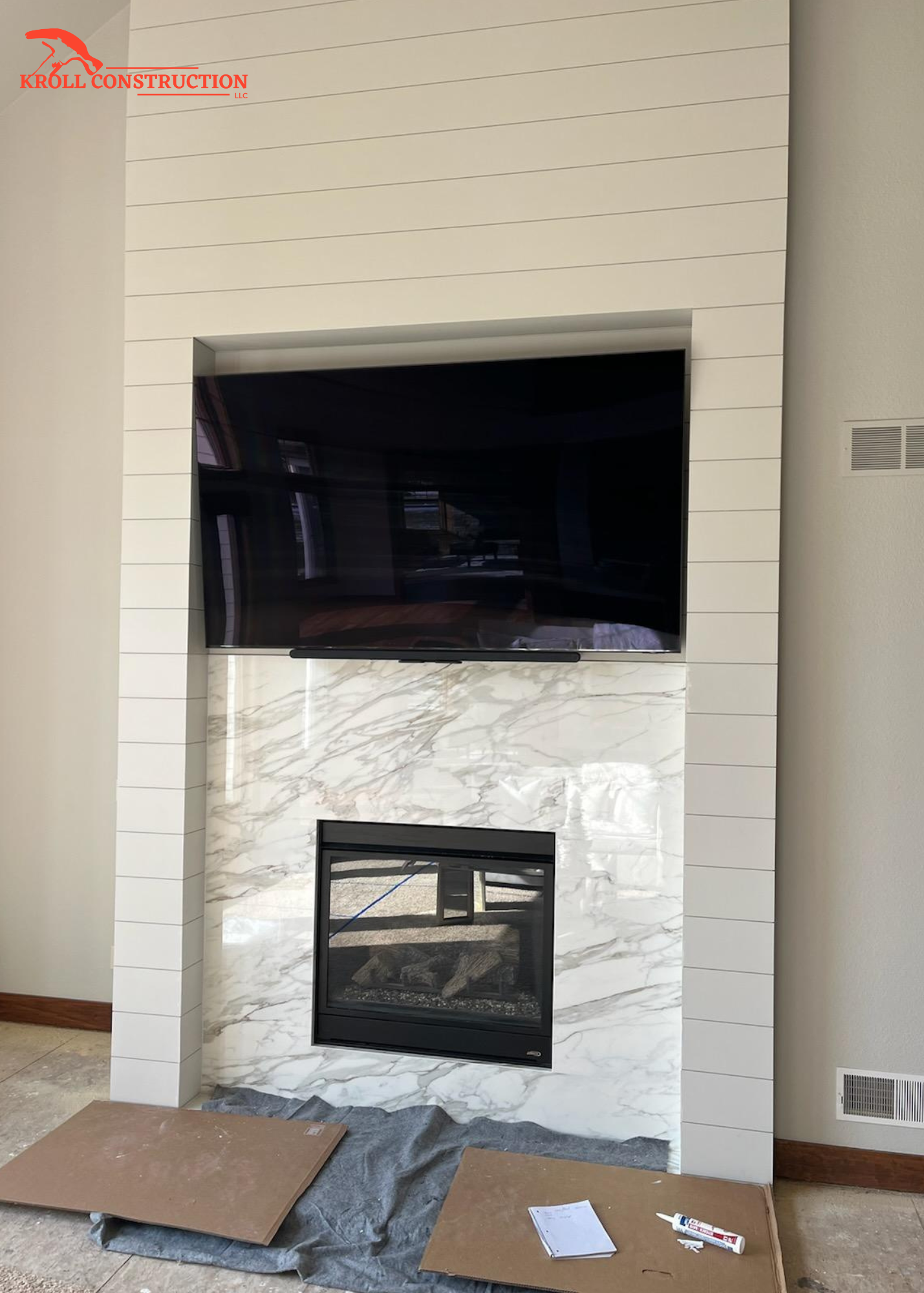 A modern fireplace with a marble surround and a black firebox, mounted on a wall with white horizontal paneling above and to the sides, in a room under construction, with tiles and tools on the floor.