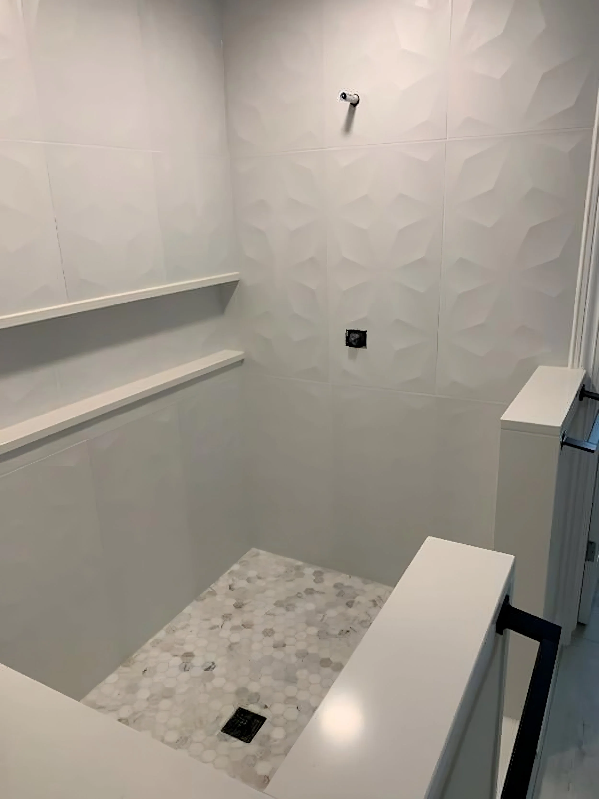 Unfinished bathroom shower area with white tiles, built-in shelves, a shower head pipe, and a floor with small hexagonal tiles.